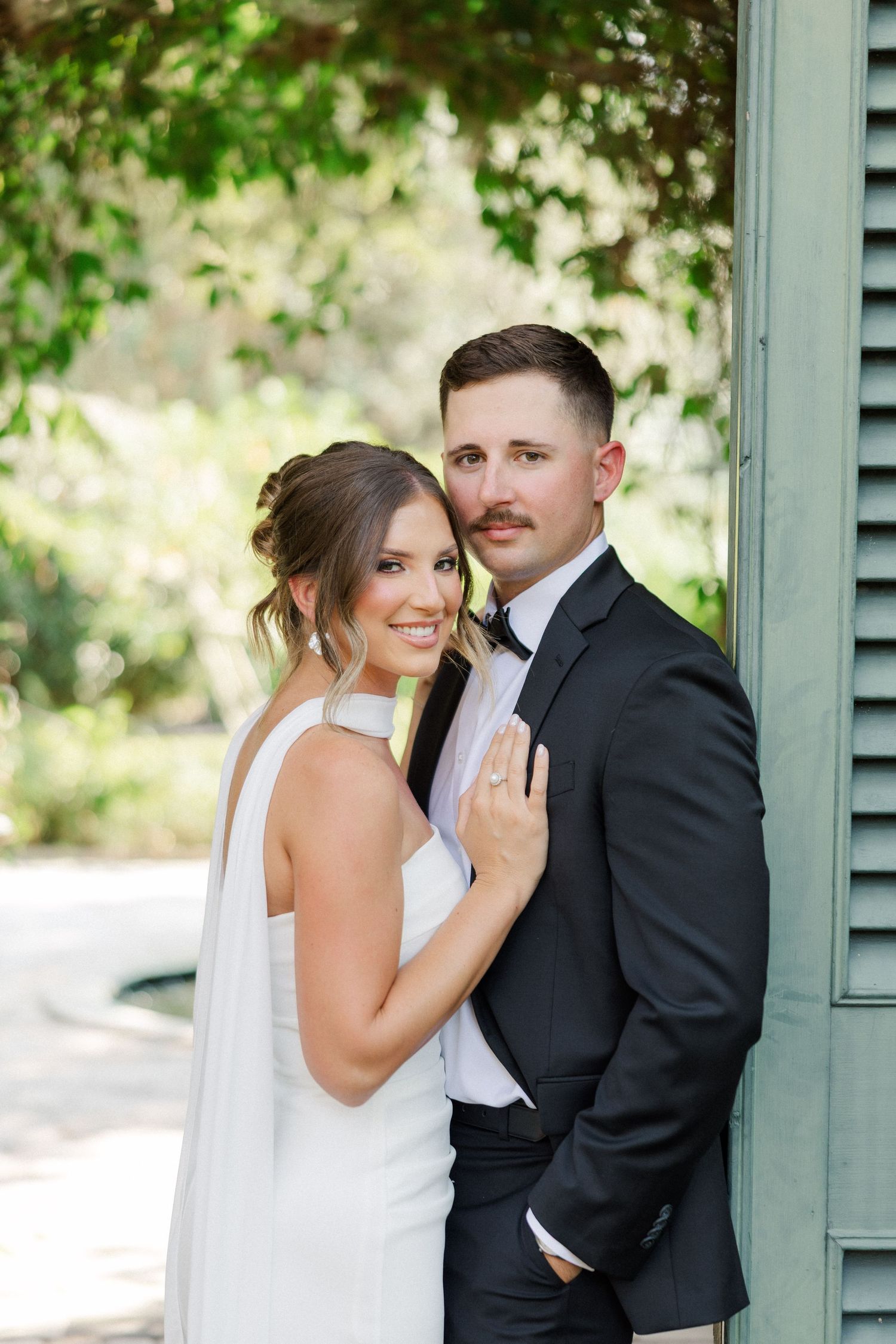 A romantic wedding portrait of a couple in formal attire posing together against a soft outdoor background.