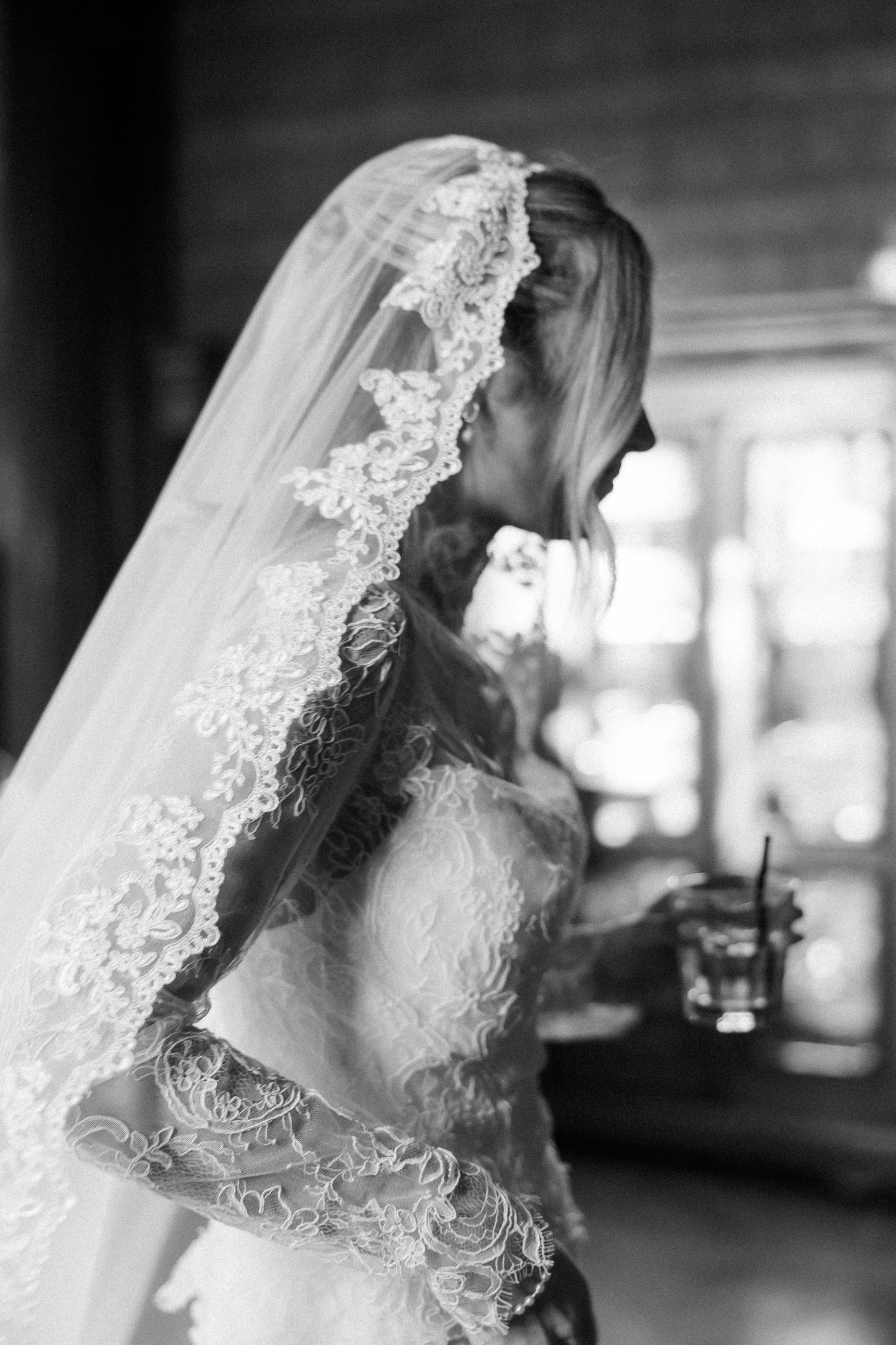 A delicate lace veil frames a bridal portrait in black and white with soft window lighting.
