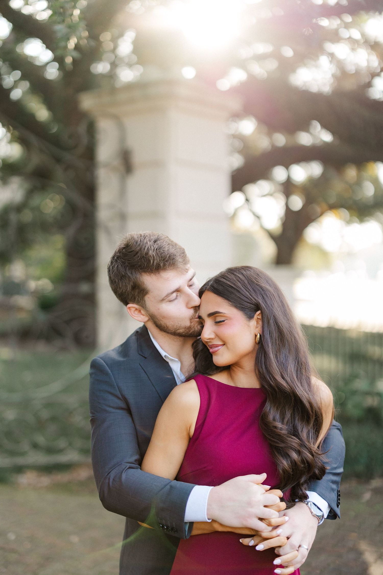 Intimate moment captured between couple in formal attire against sunlit garden backdrop.