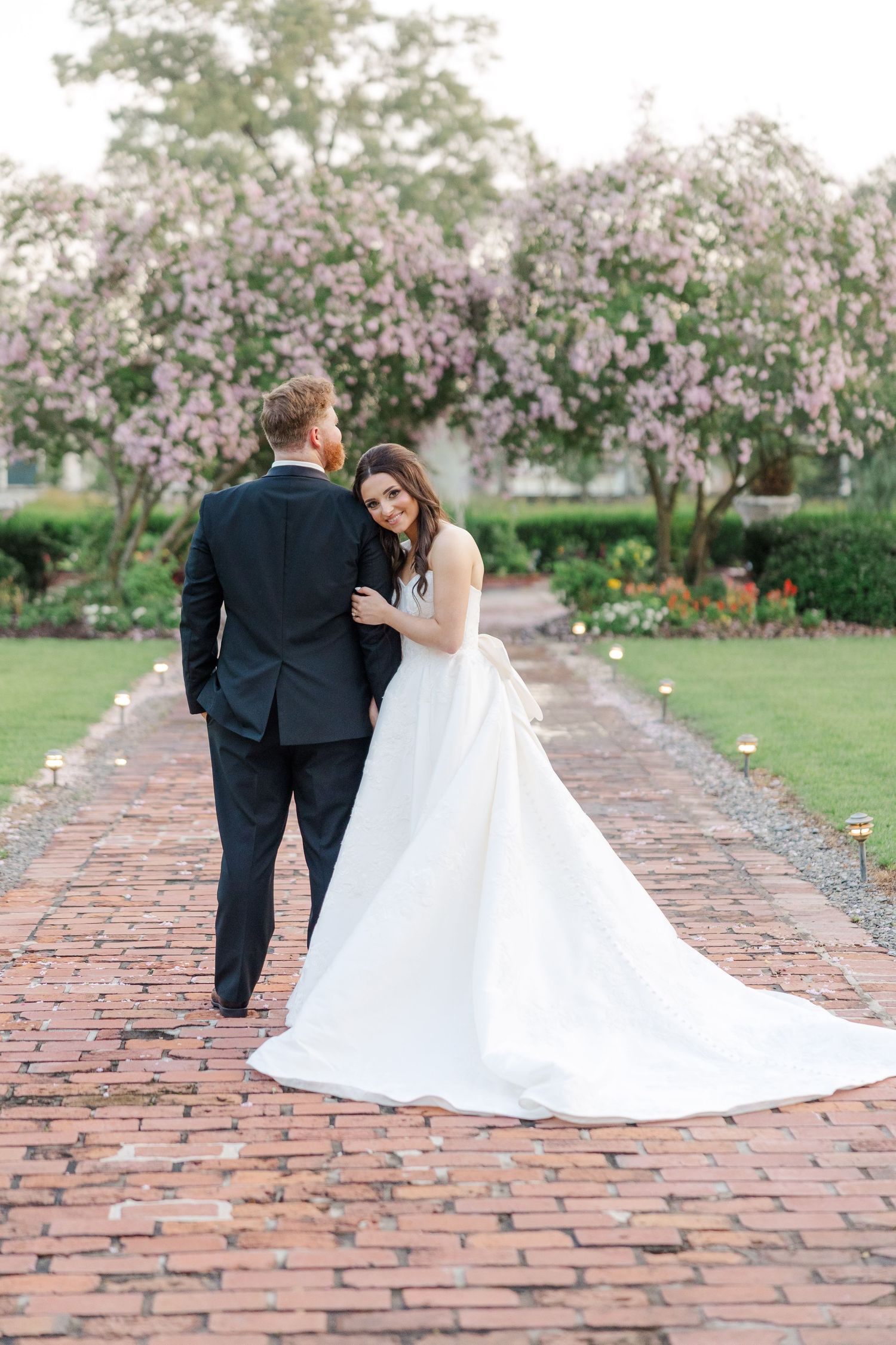 Couple in wedding attire walking along brick pathway surrounded by blooming trees.