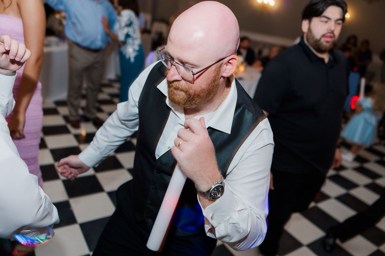 A guest with red hair and beard dances enthusiastically on a checkered dance floor at a wedding reception.
