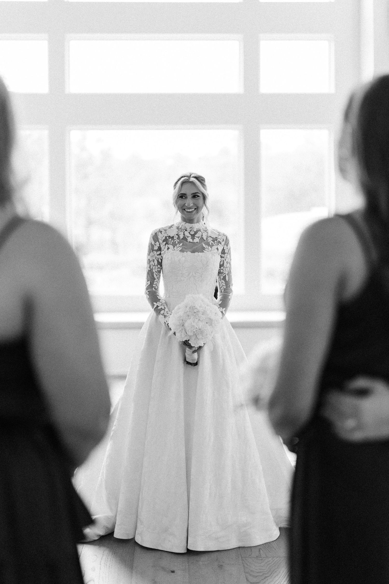 A delicate white lace wedding dress is displayed in a bright room with large windows.