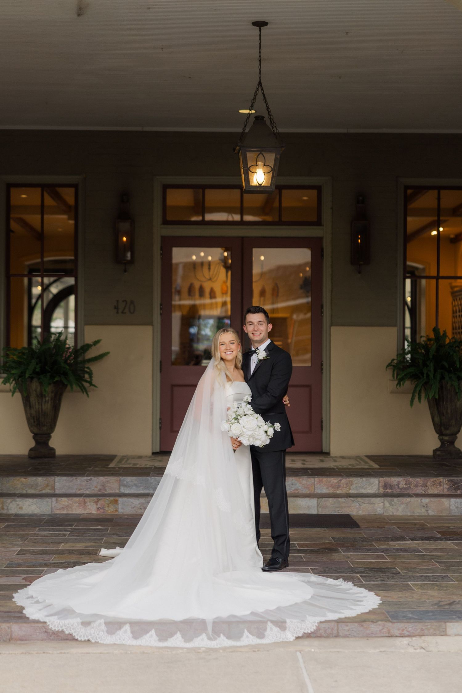 A wedding couple poses on an elegant porch with hanging lanterns and potted plants, featuring a long flowing white train.