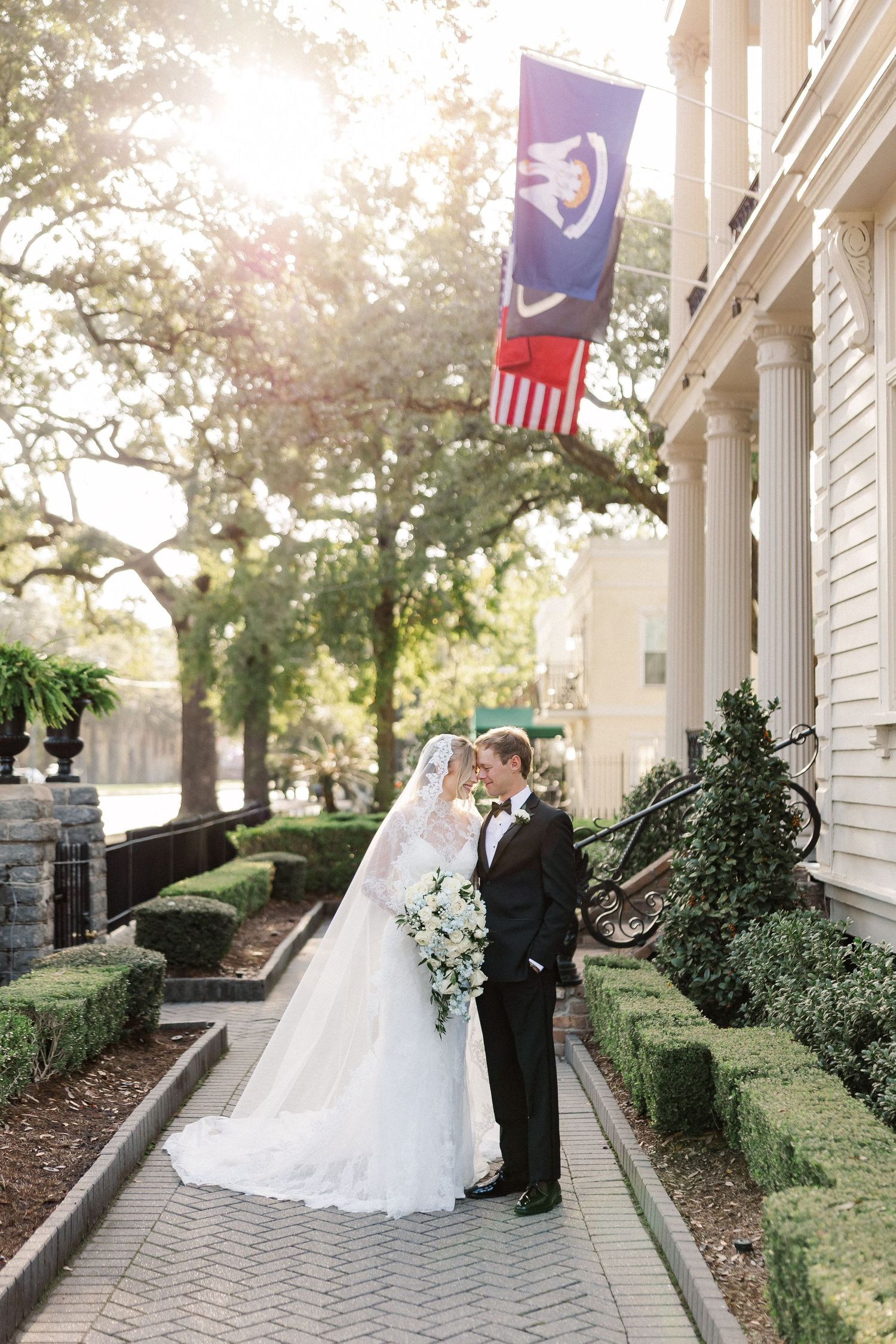 Elegant wedding couple poses along white columned porch with hanging flags and manicured hedges in afternoon light.