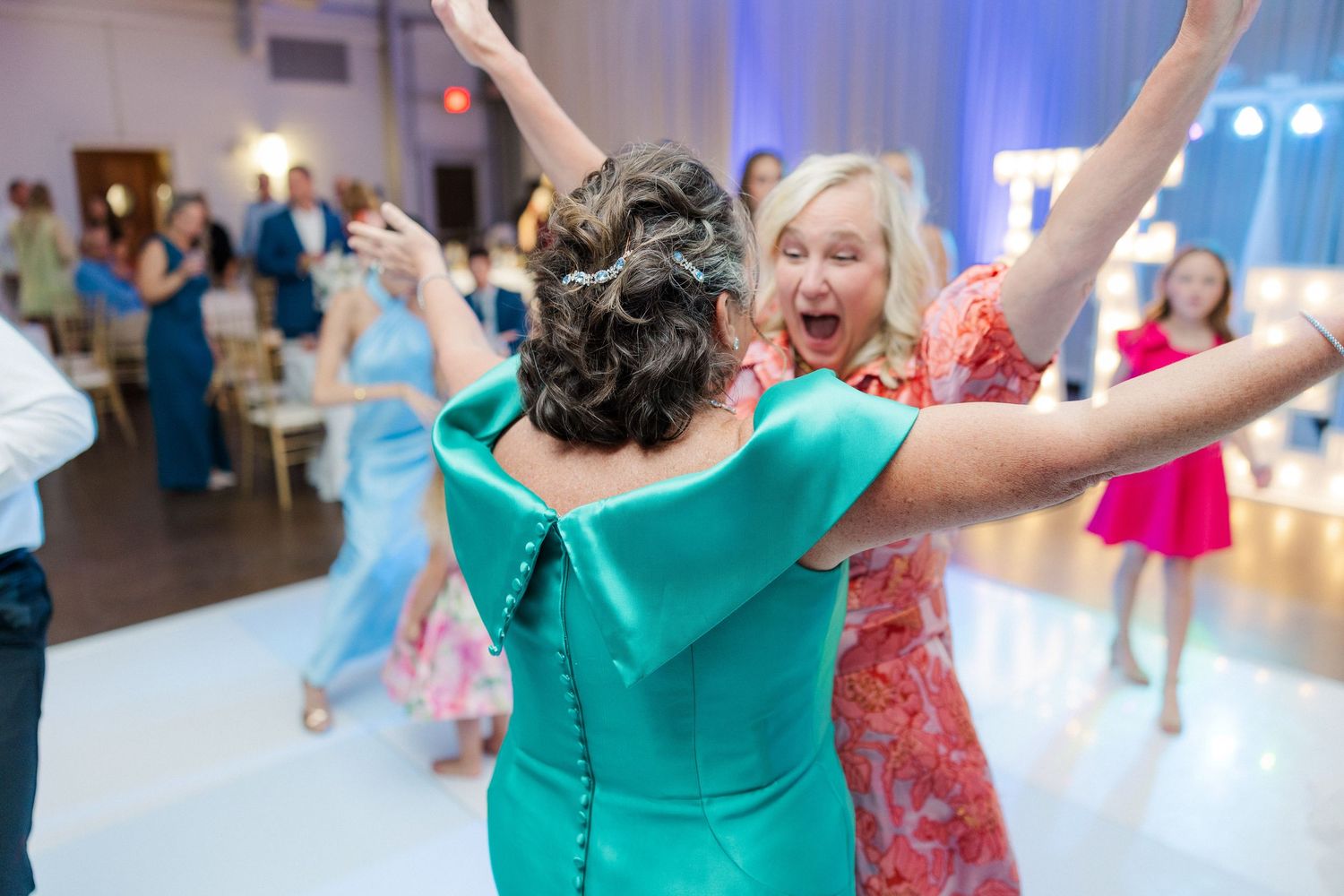 Wedding guests in colorful dresses dance enthusiastically at the reception.