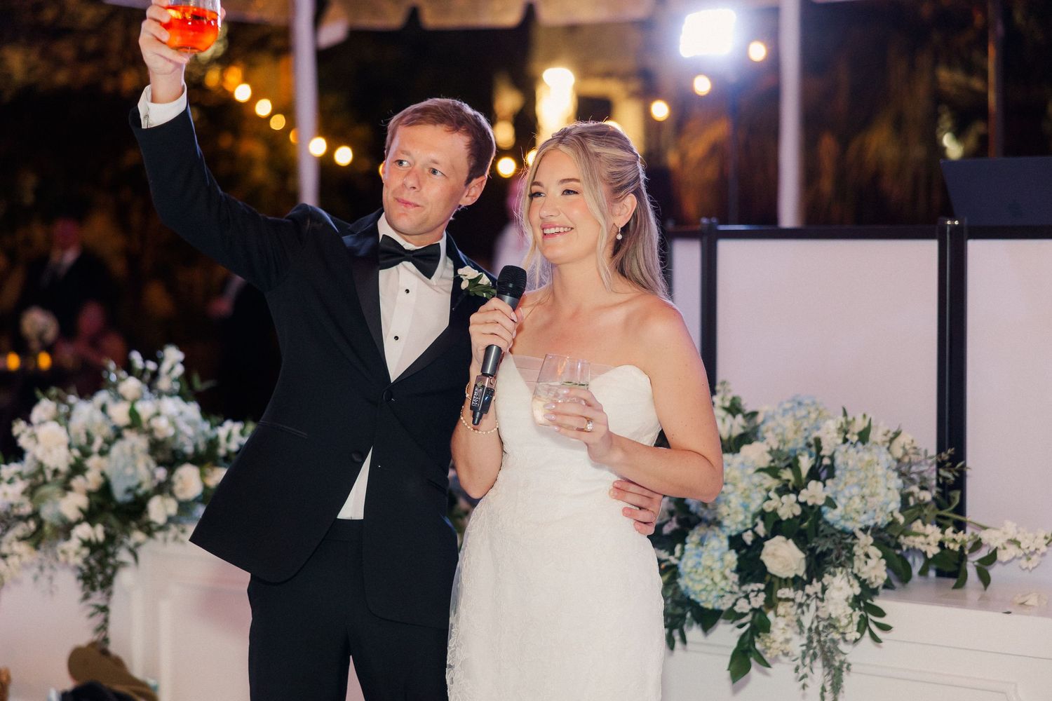 Wedding reception celebration with couple toasting under string lights and floral decorations.