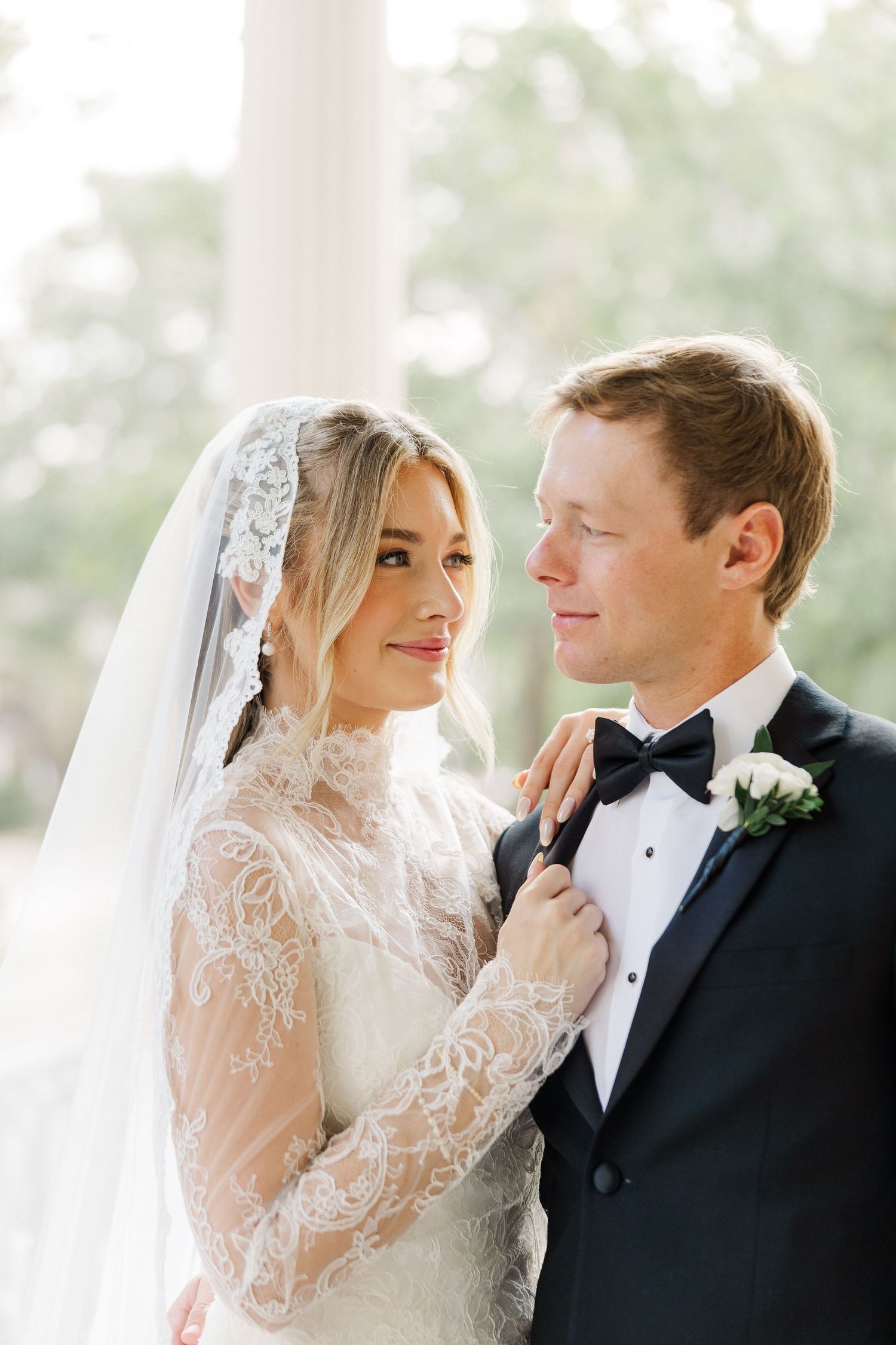 Elegant wedding couple in formal attire sharing an intimate moment in front of bright windows.