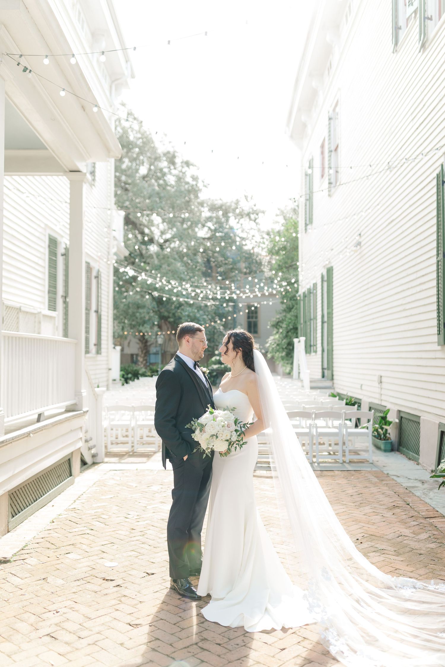 Newlyweds share an intimate moment beneath string lights on a brick courtyard between white buildings.