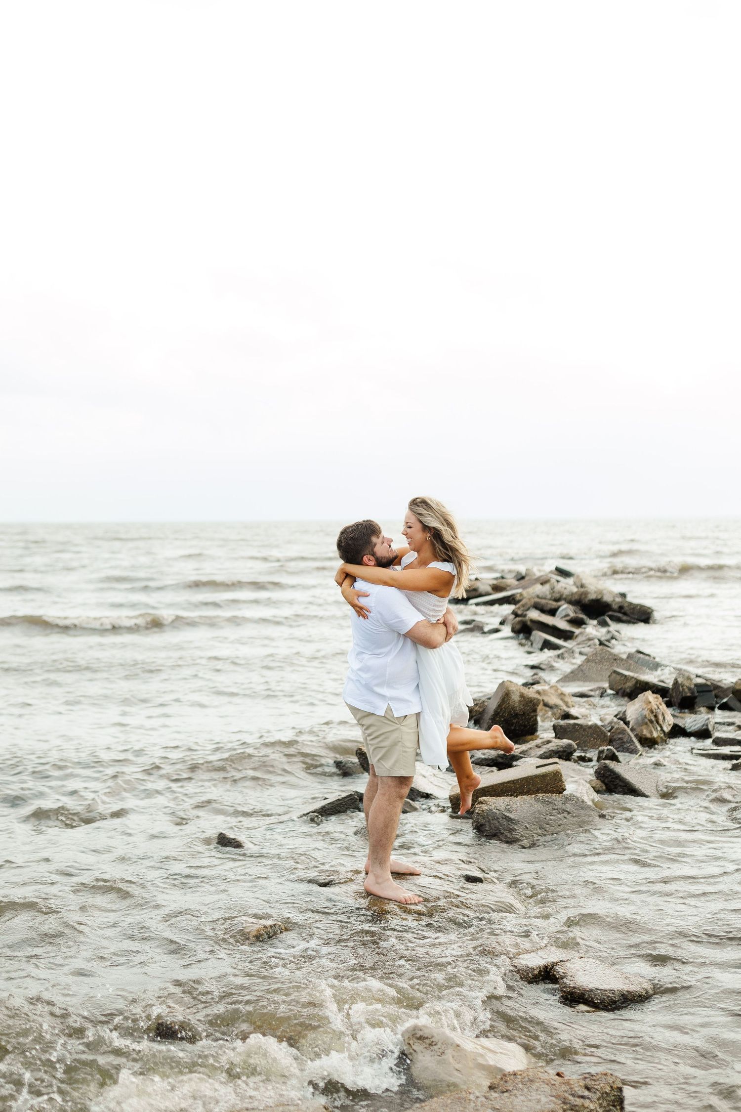 Couple embraces romantically on rocky beach with crashing waves and overcast sky in the background.