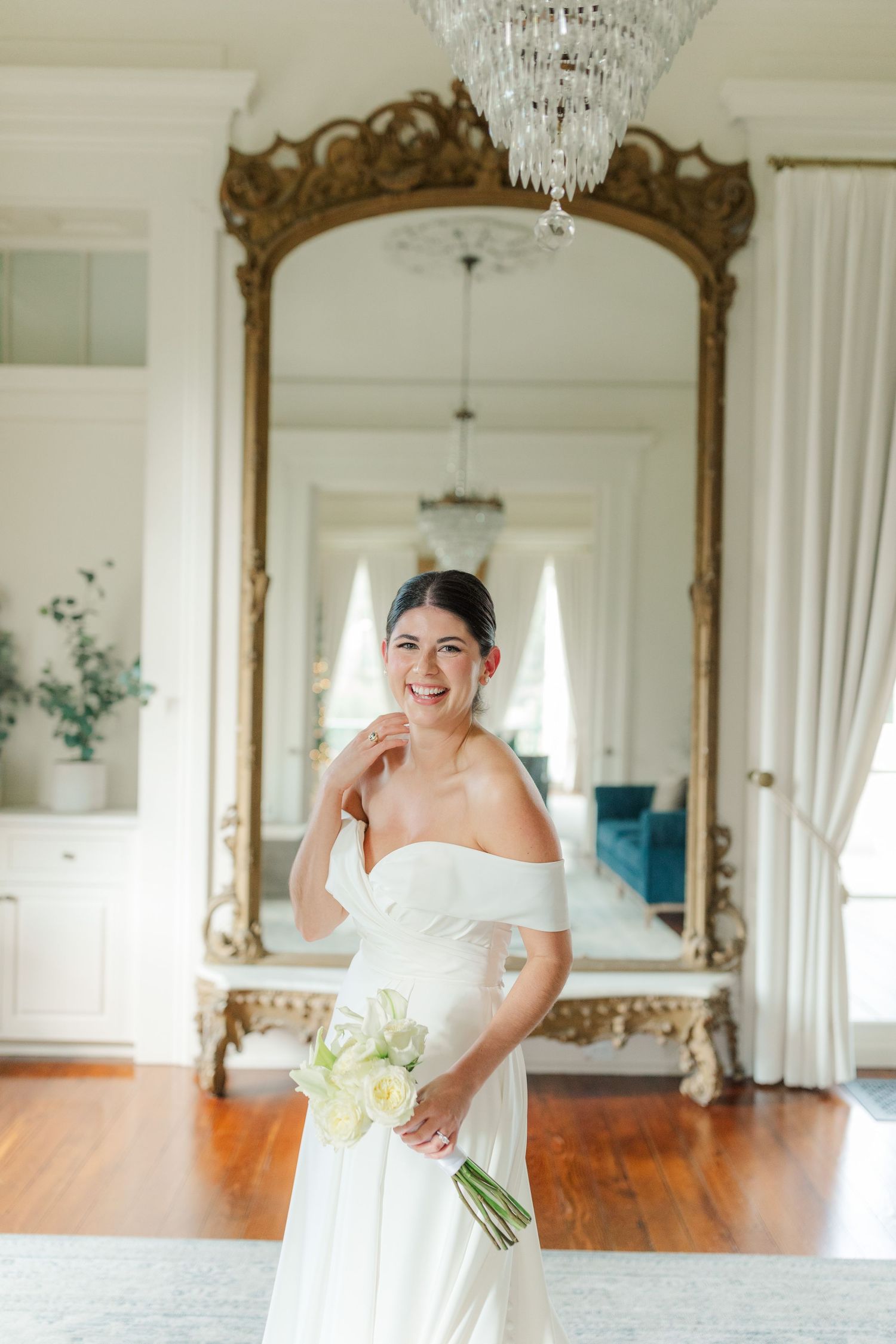 Elegant bride in off-shoulder white gown poses in front of ornate gold mirror with crystal chandelier in bright room.