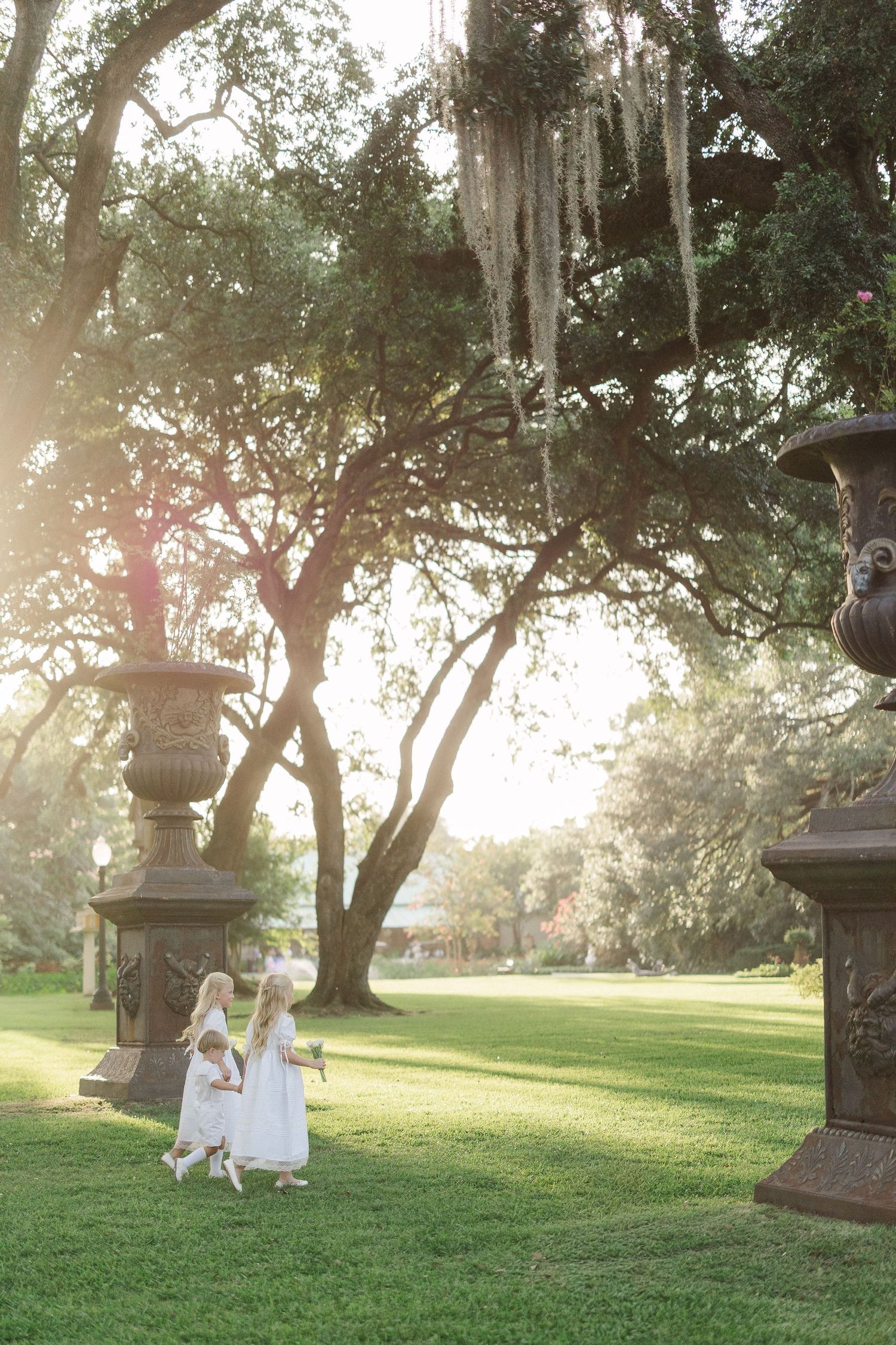 Two flower girls in white dresses walk across a sunlit lawn beneath moss-draped oak trees at a garden wedding.