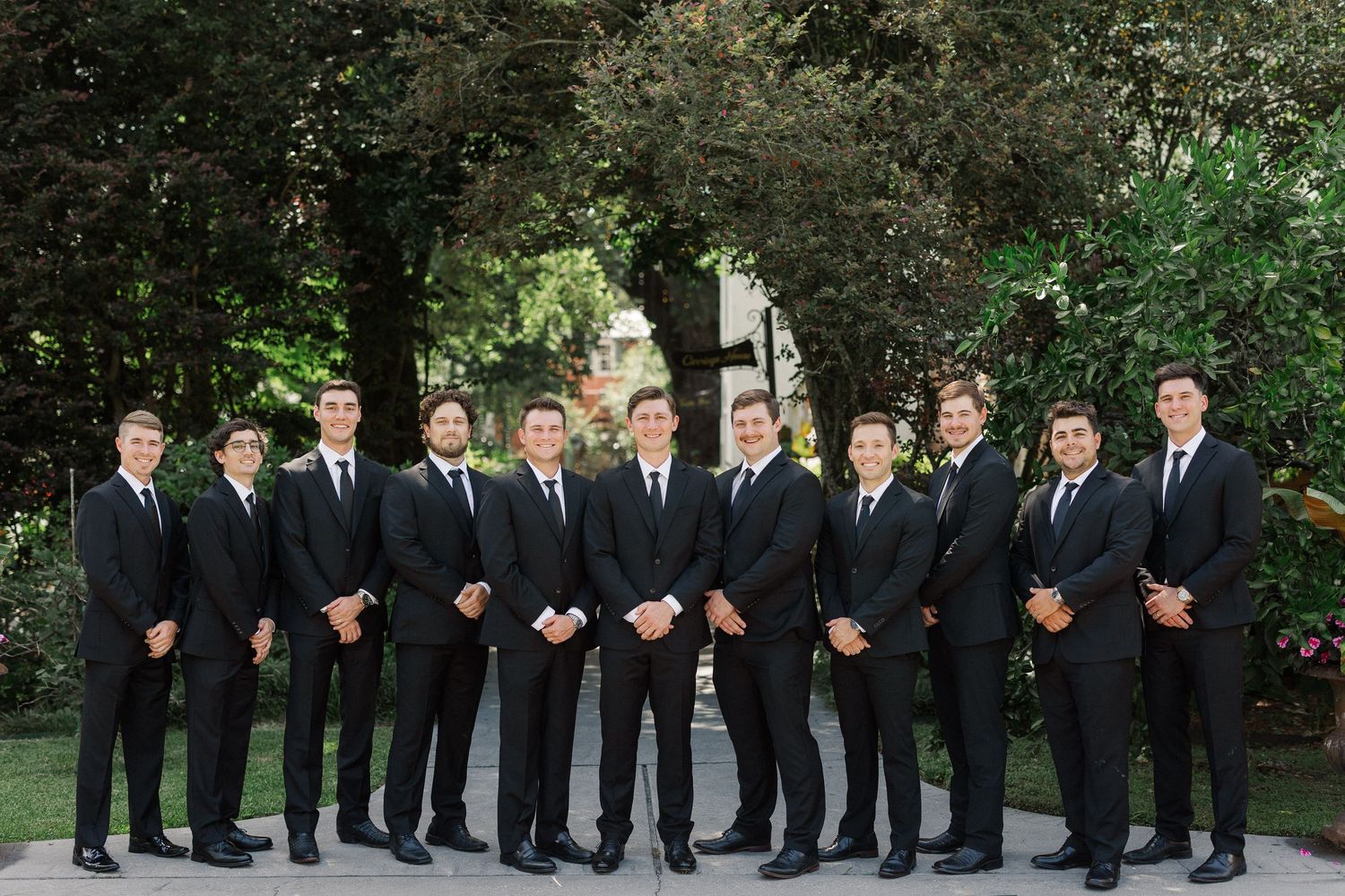 Eleven groomsmen in matching black suits pose in a row outdoors with lush green trees in the background.