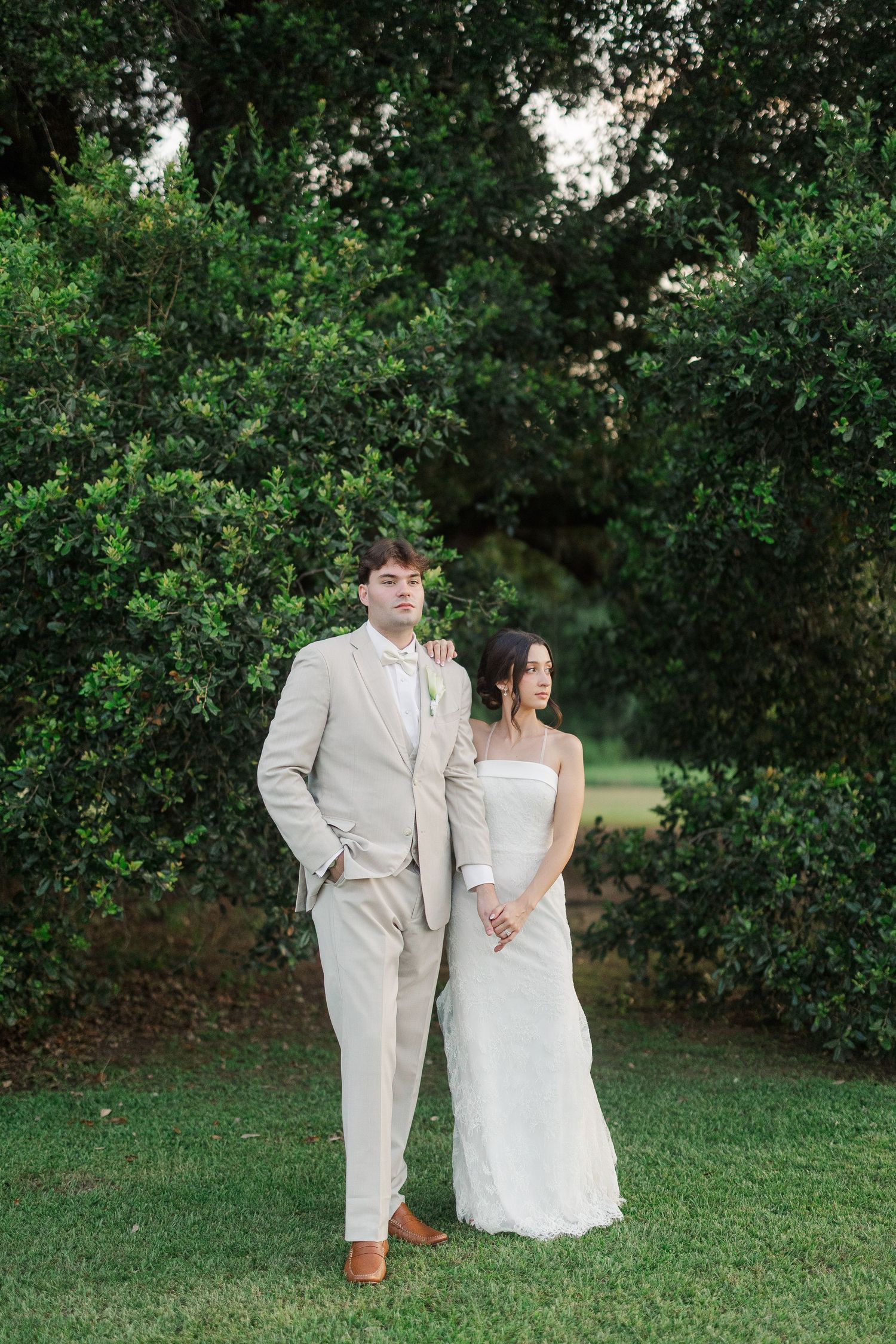 Bride and groom pose together in front of lush green hedges, groom in beige suit, bride in strapless white gown.