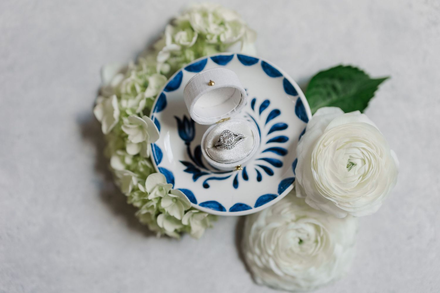 Wedding rings in a blue and white ceramic dish surrounded by white hydrangeas and ranunculus flowers on a gray surface.