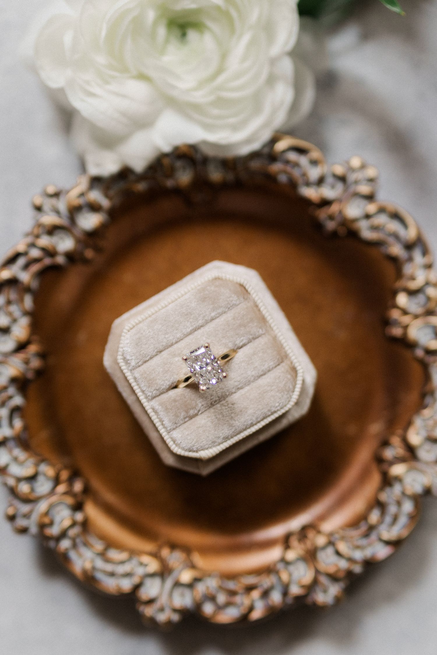 Diamond solitaire engagement ring in a velvet ring box on an ornate gold tray with white ranunculus flowers.