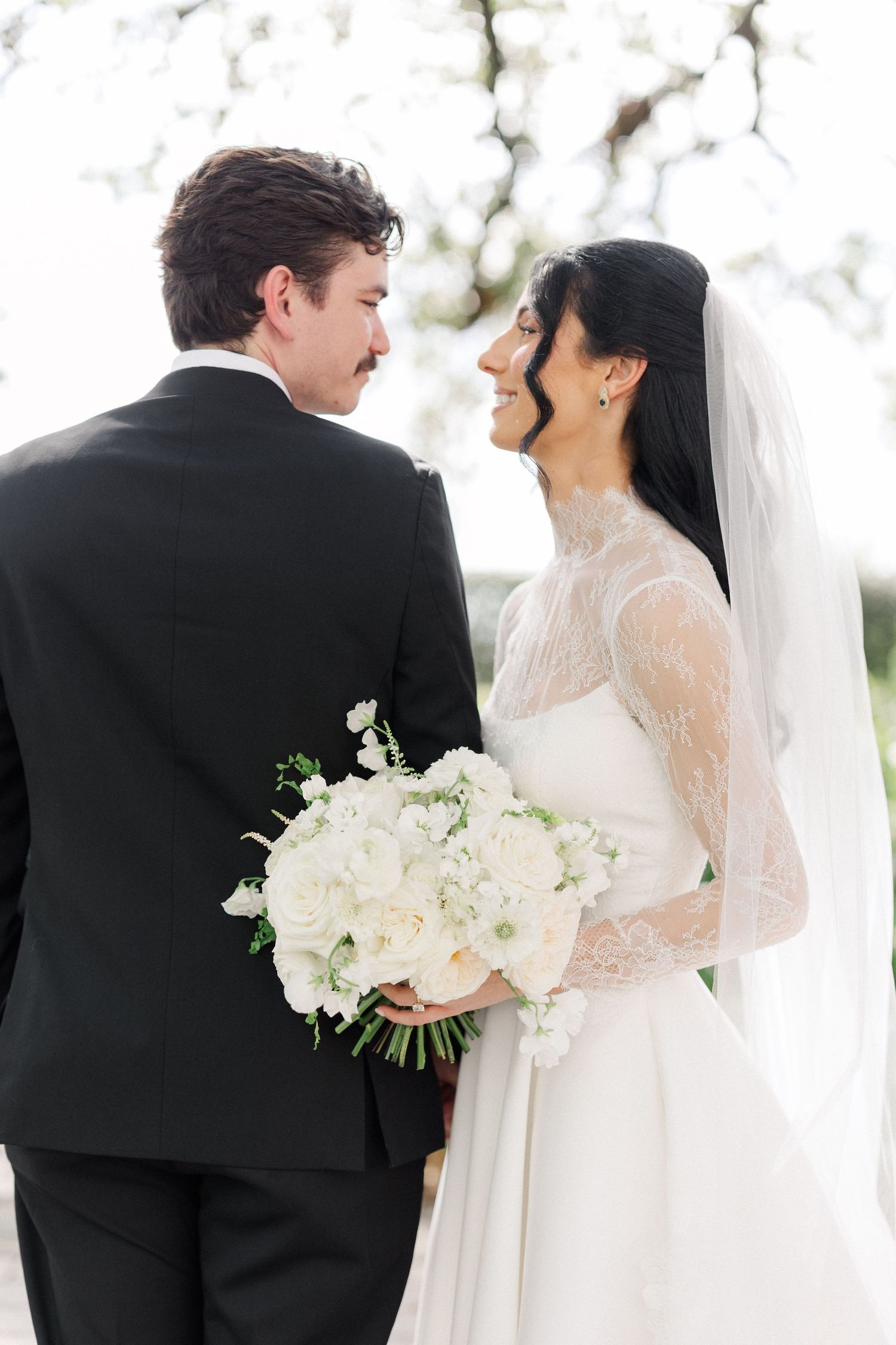 Bride in lace veil smiles at groom holding white rose bouquet outdoors under blooming trees.
