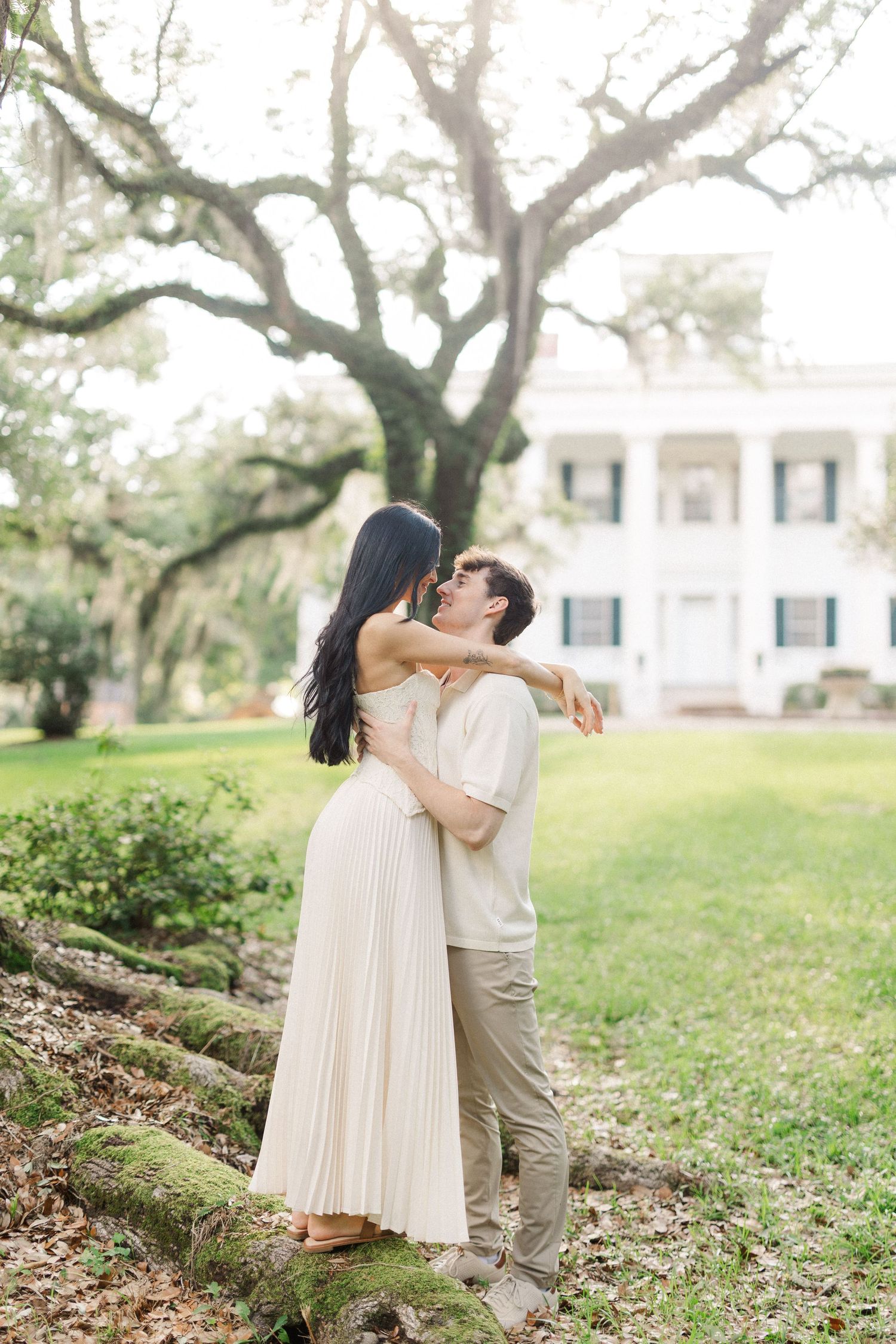 Couple embracing and kissing outdoors near a grand white Southern plantation home with moss-covered oak trees.