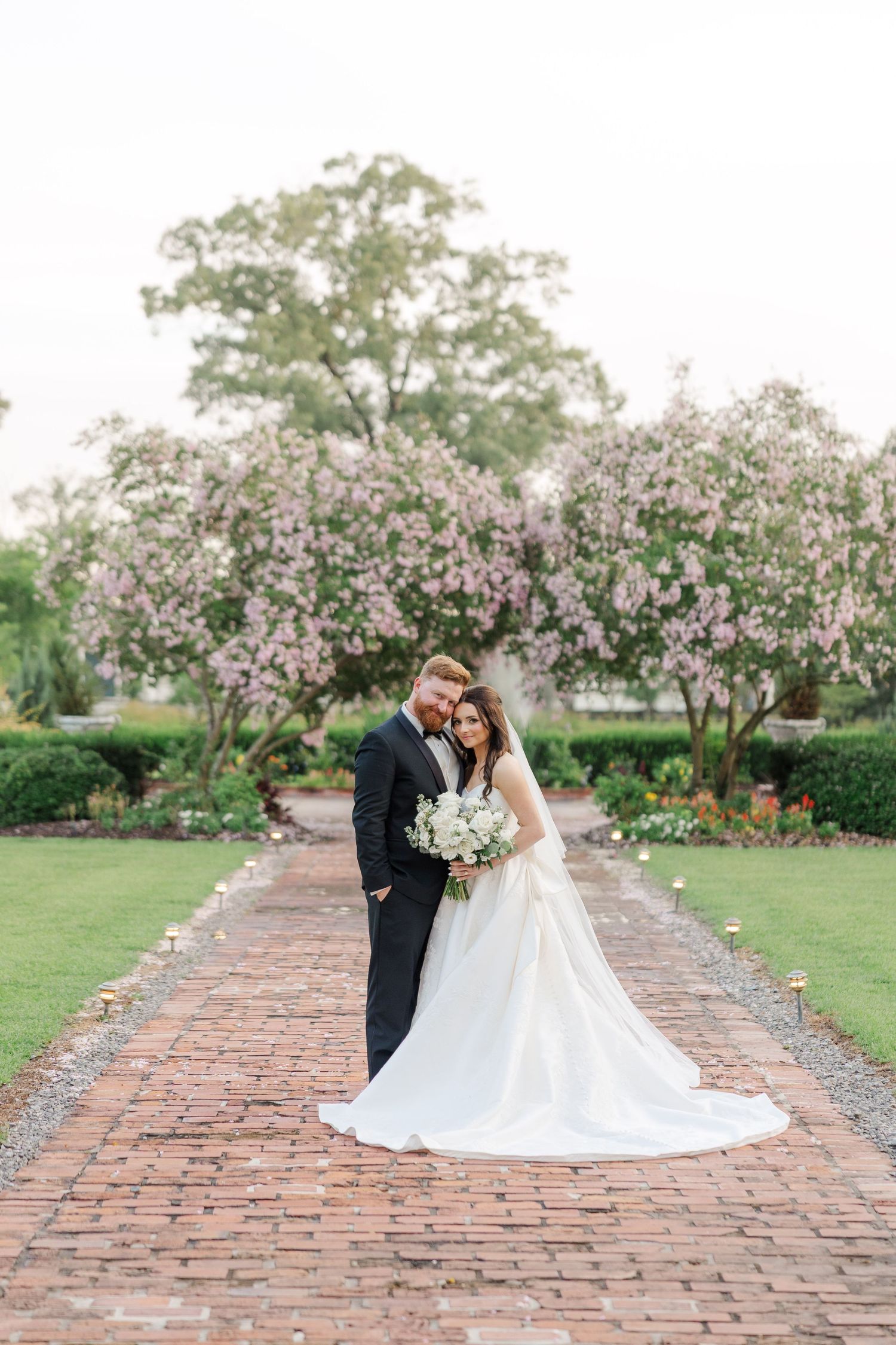 Newlyweds pose on a brick pathway under blooming cherry trees in a garden setting.