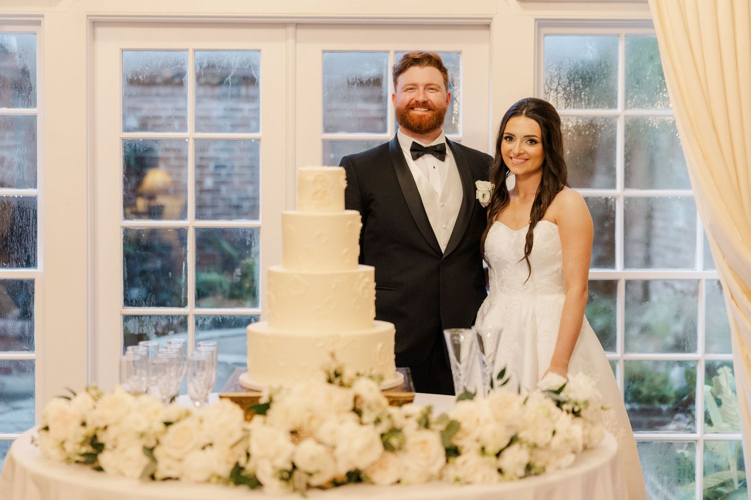 Elegant wedding reception venue with large windows, white curtains, and floral centerpieces on tables.