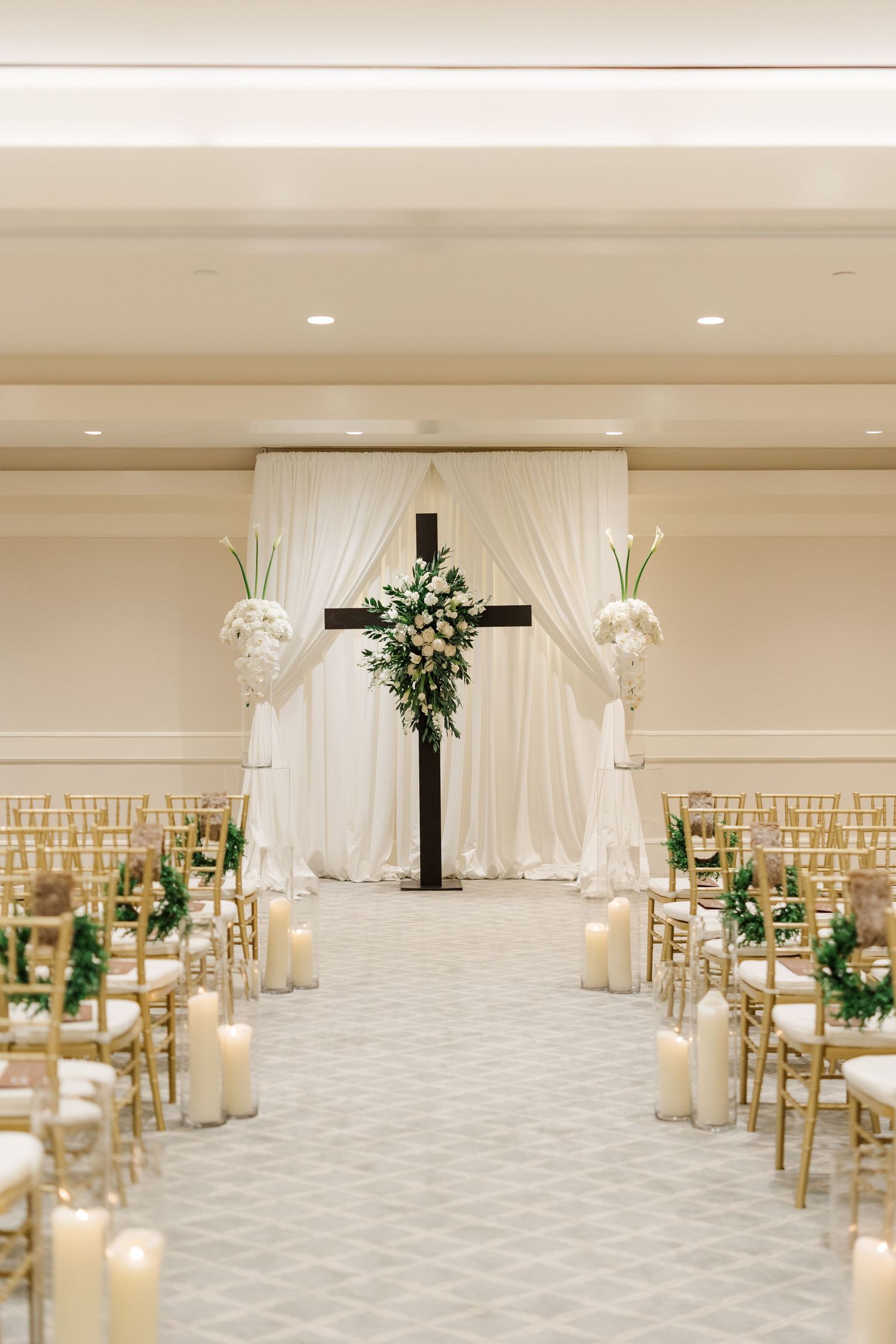 Elegant wedding ceremony setup with cross, white draping, and gold chairs lined with candles.
