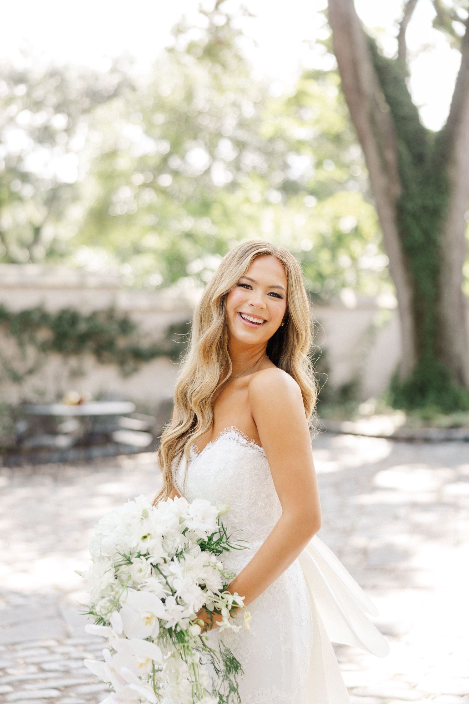 A bride in a white strapless wedding dress poses outdoors in natural sunlight holding a white floral bouquet.