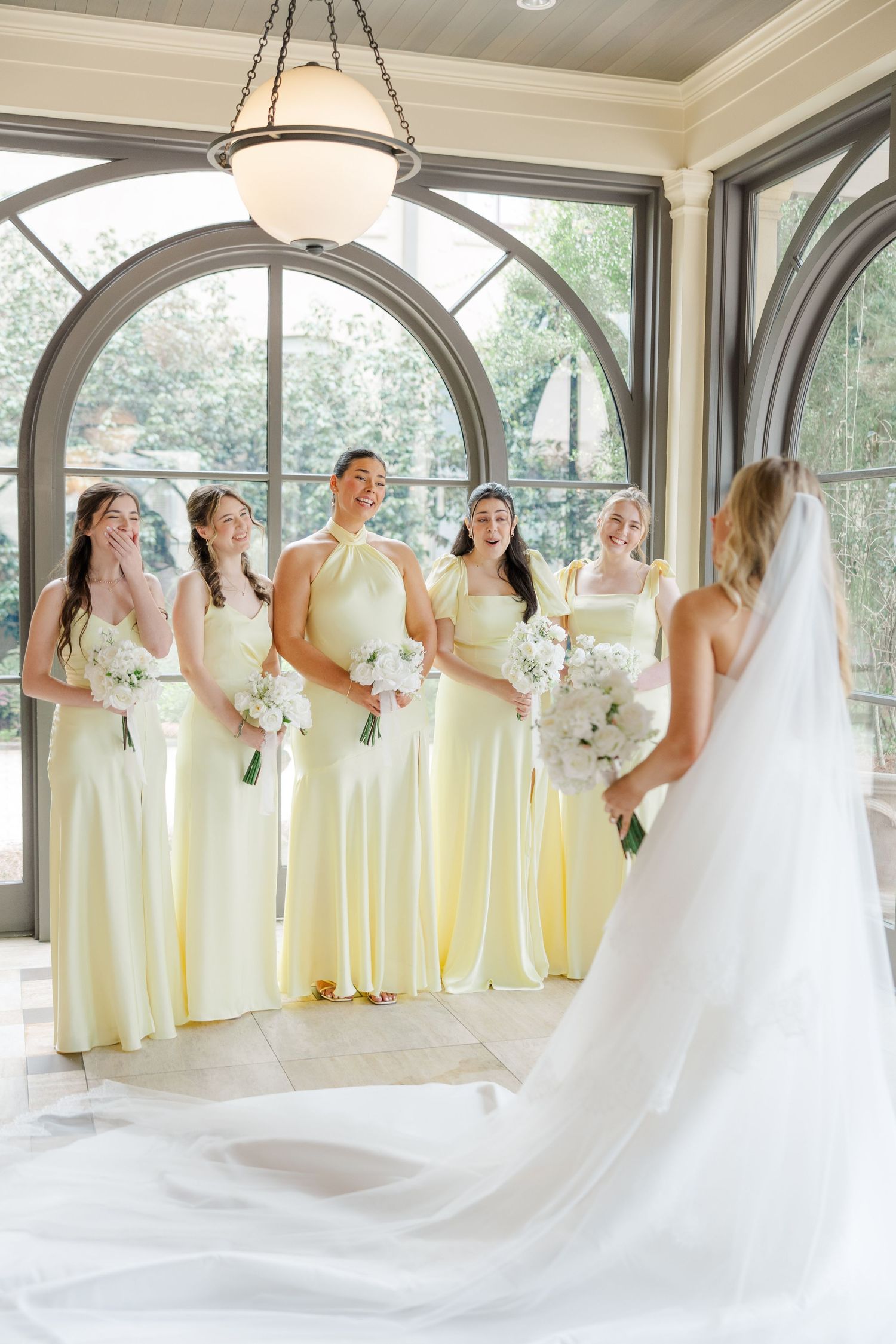 A bridal party in pale yellow dresses poses in an elegant sunlit room with arched windows and white floral bouquets.