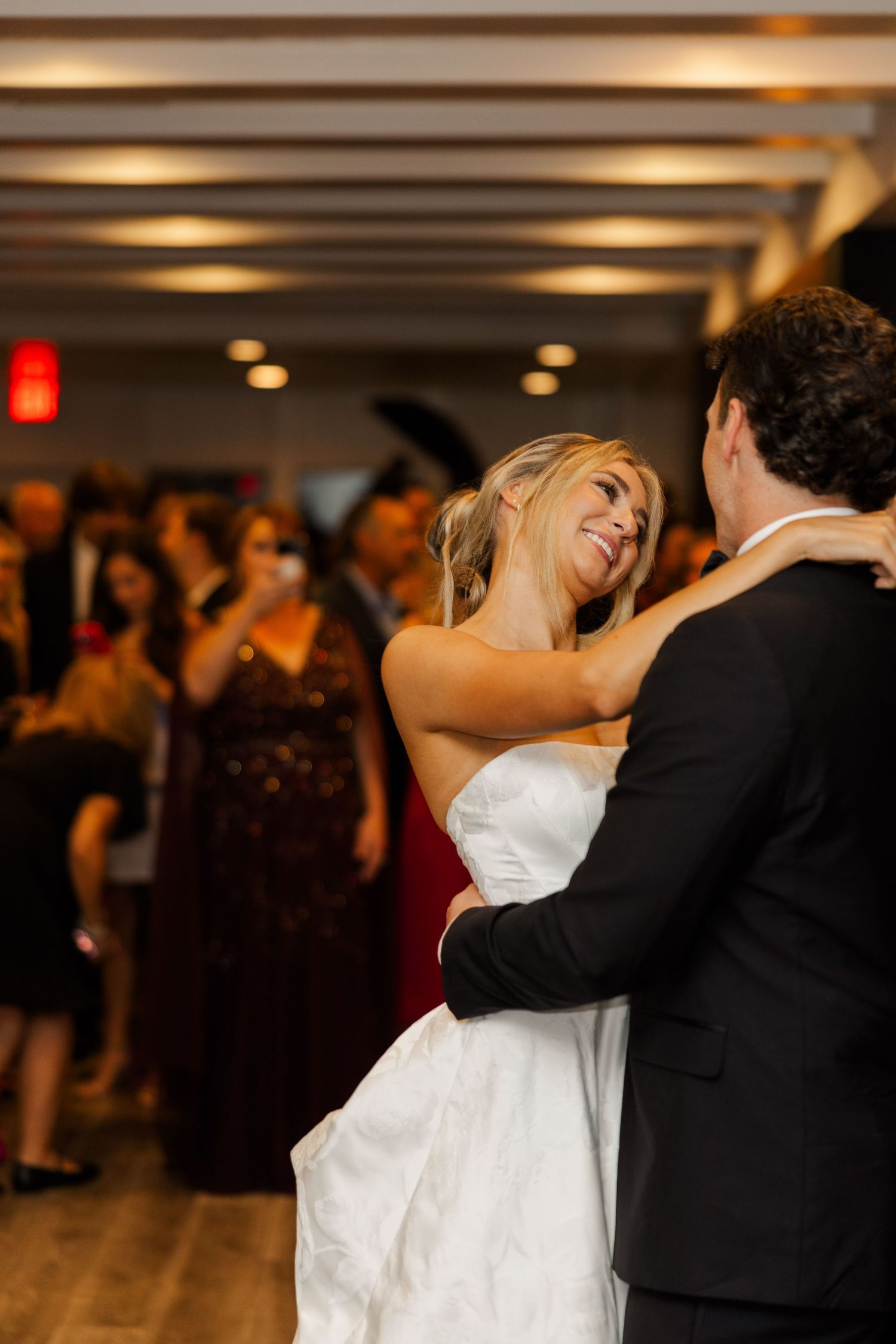 Wedding couple dances surrounded by guests in a warmly lit reception venue.