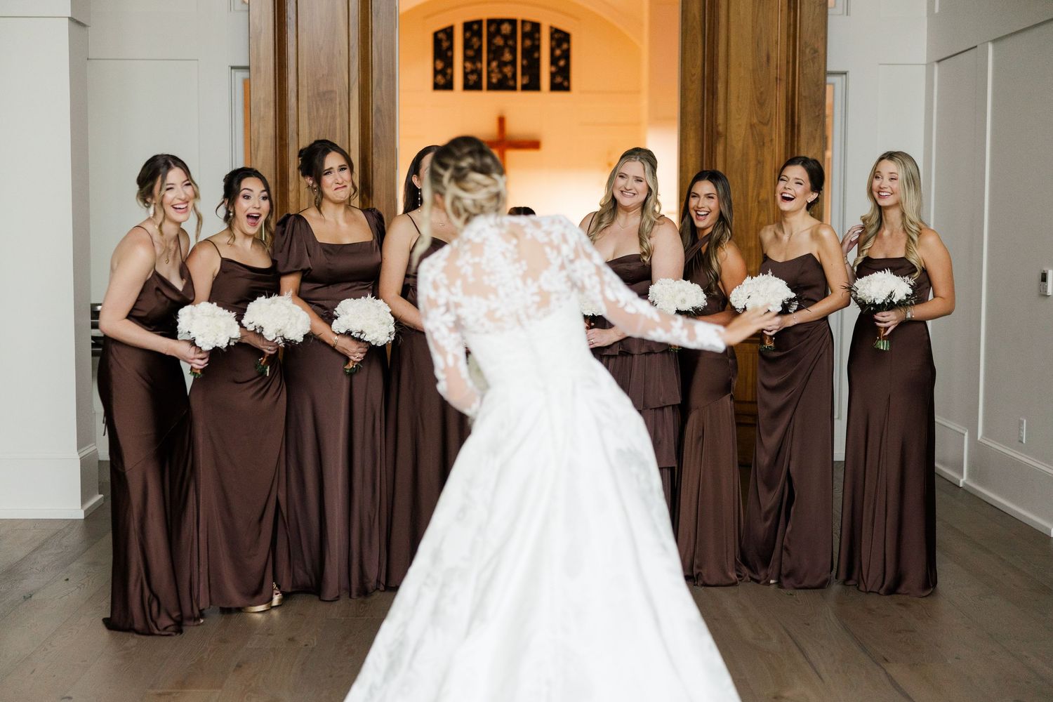 A bridal party in chocolate brown dresses and a bride in white gather in an elegant hallway for a wedding photo.