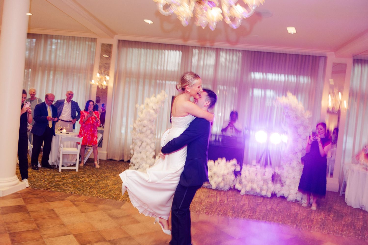 Wedding reception venue decorated with pink lighting and chandeliers as guests dance on the ballroom floor.