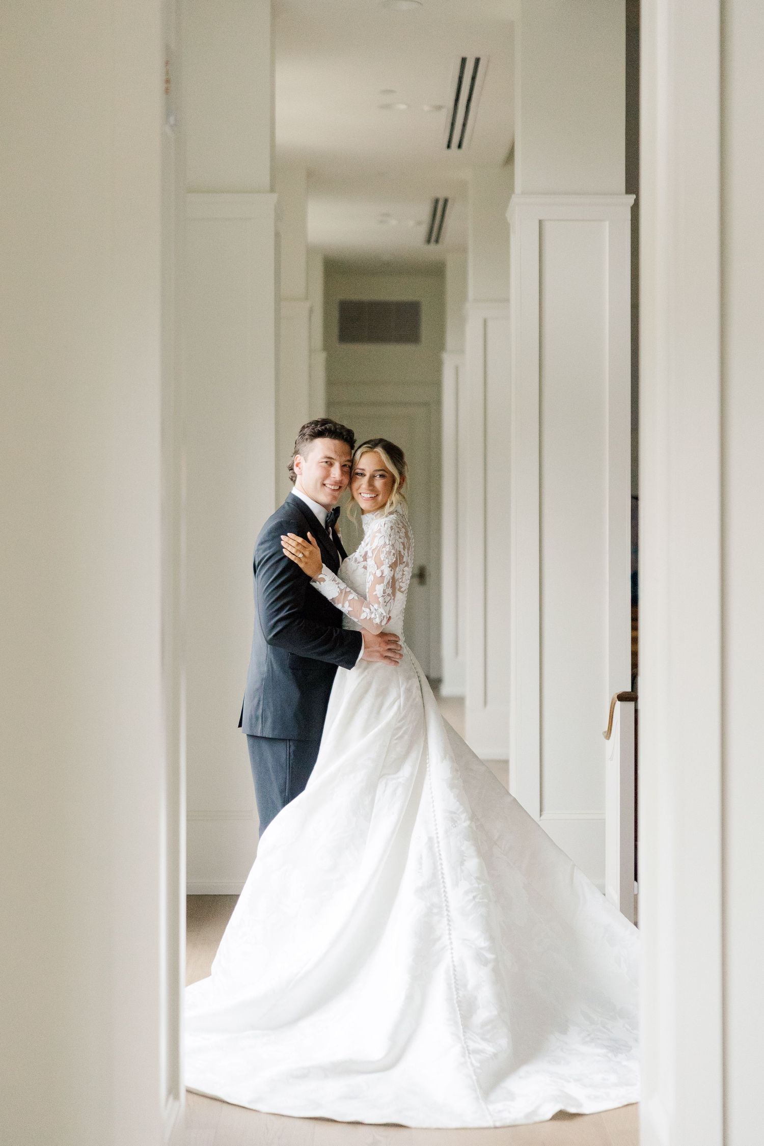 A bride in a flowing white gown embraces her partner in an elegant white corridor.