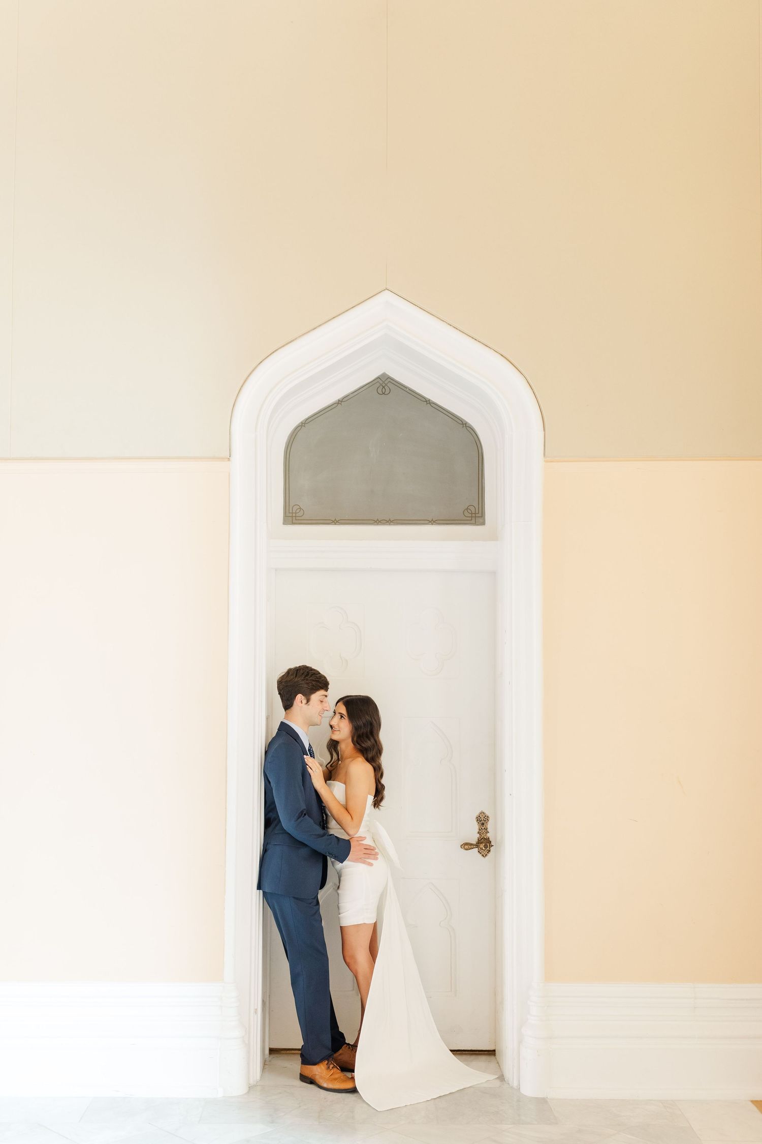 A couple in wedding attire embrace in an elegant white arched doorway with cream-colored walls.
