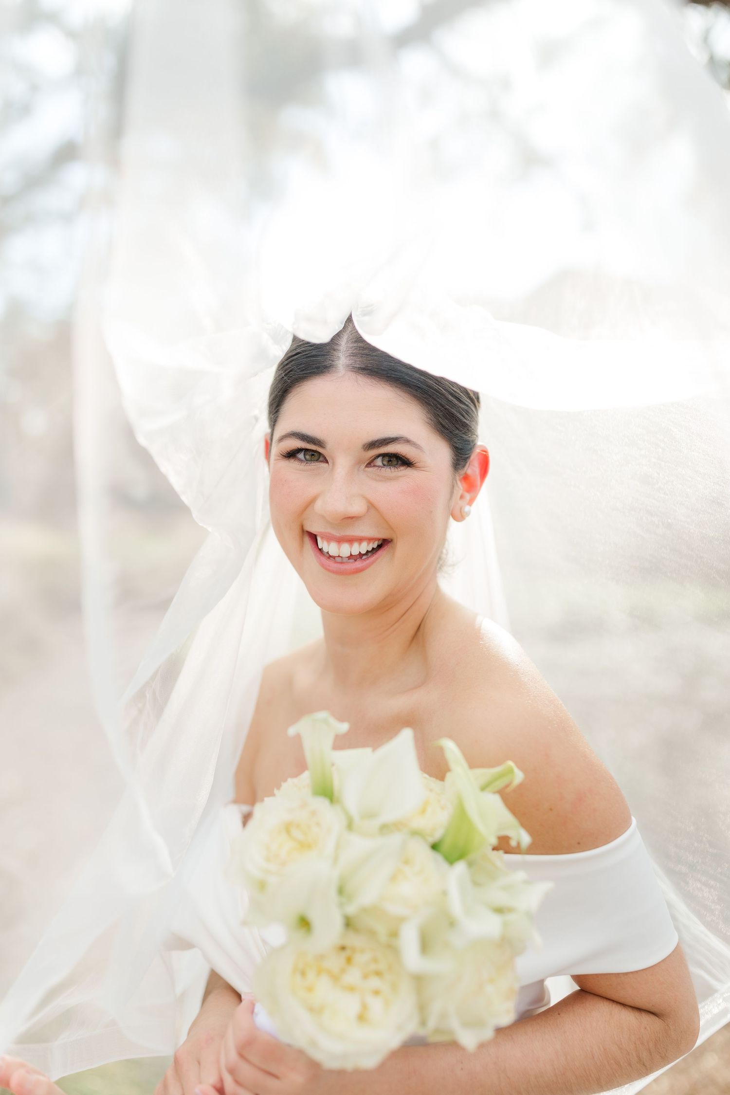 A joyful bride holds a white rose bouquet while wearing an off-shoulder wedding dress and delicate veil.
