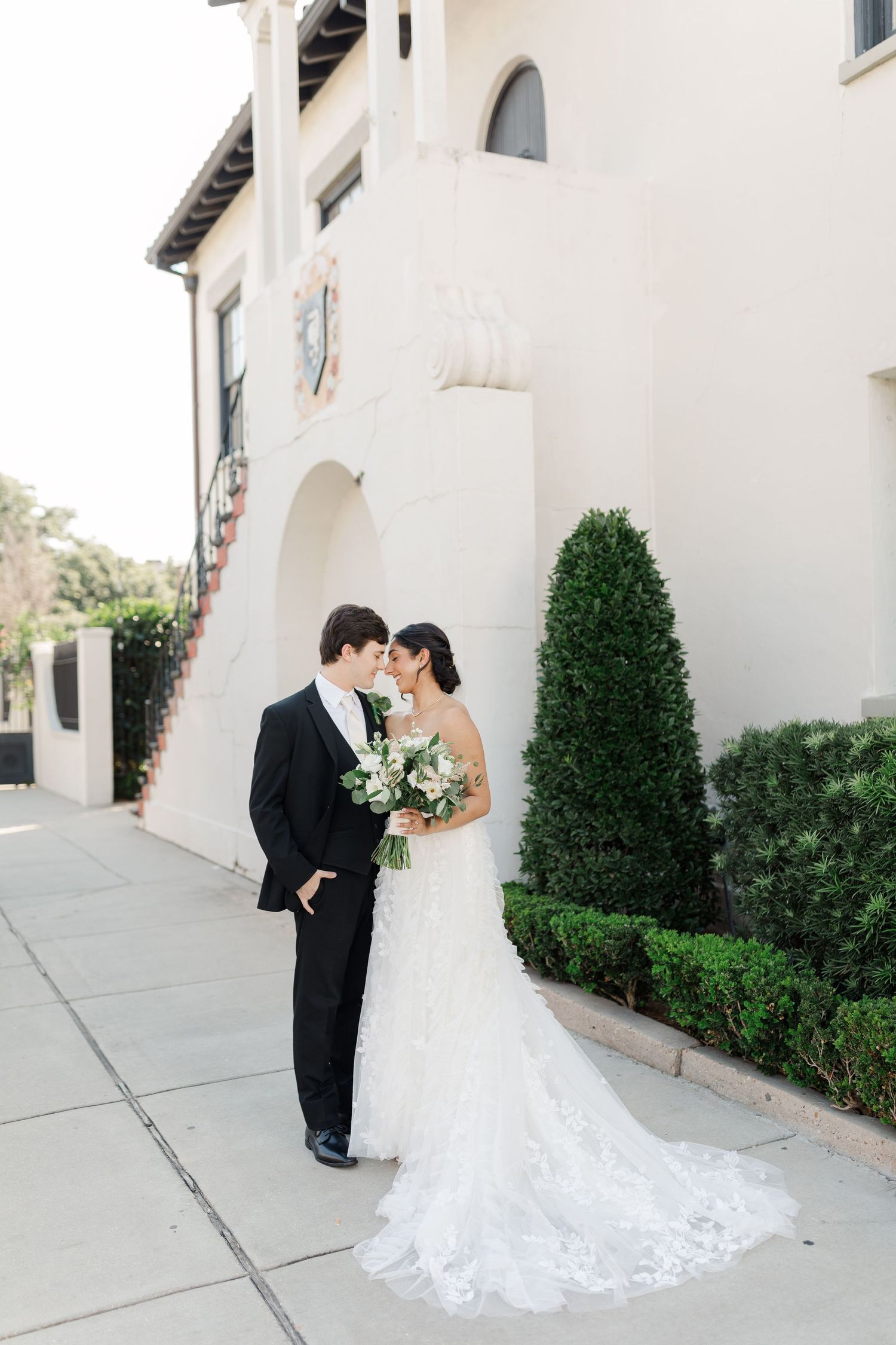 Wedding couple poses against Mediterranean-style building with topiary bushes.