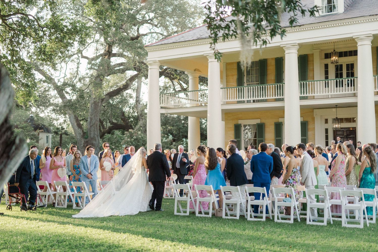 Outdoor wedding ceremony on lush lawn before grand white plantation home with guests seated in white chairs.