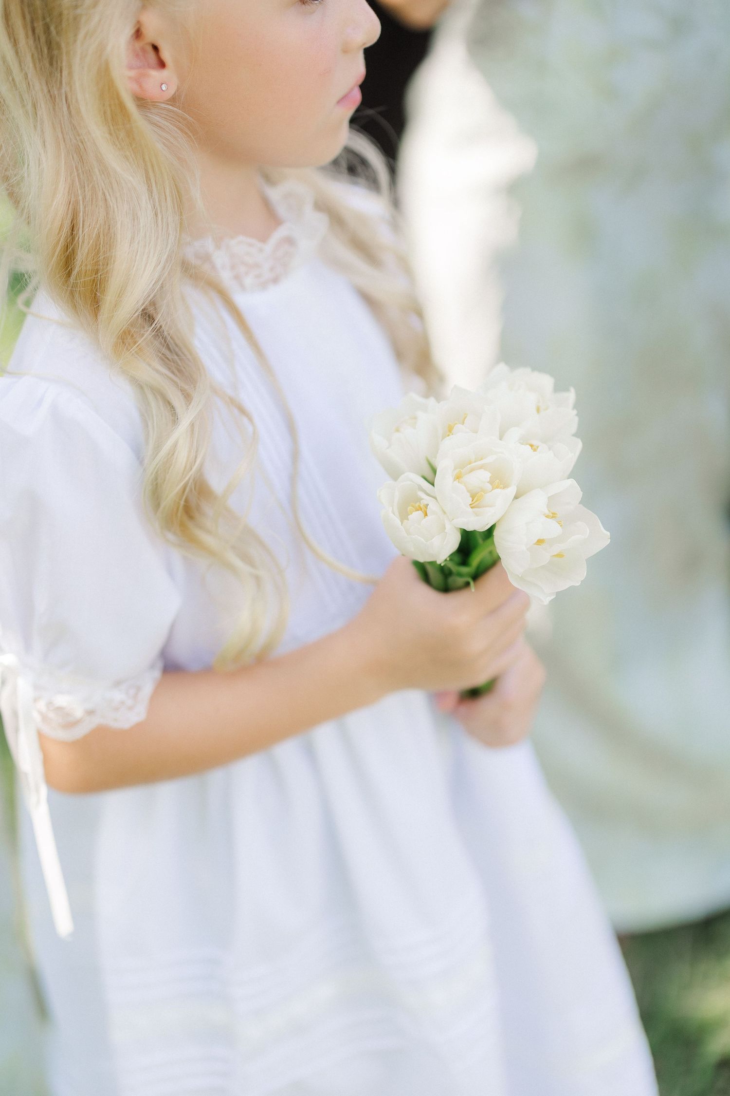 Young flower girl in white lace dress holding bouquet of white tulips at outdoor wedding.