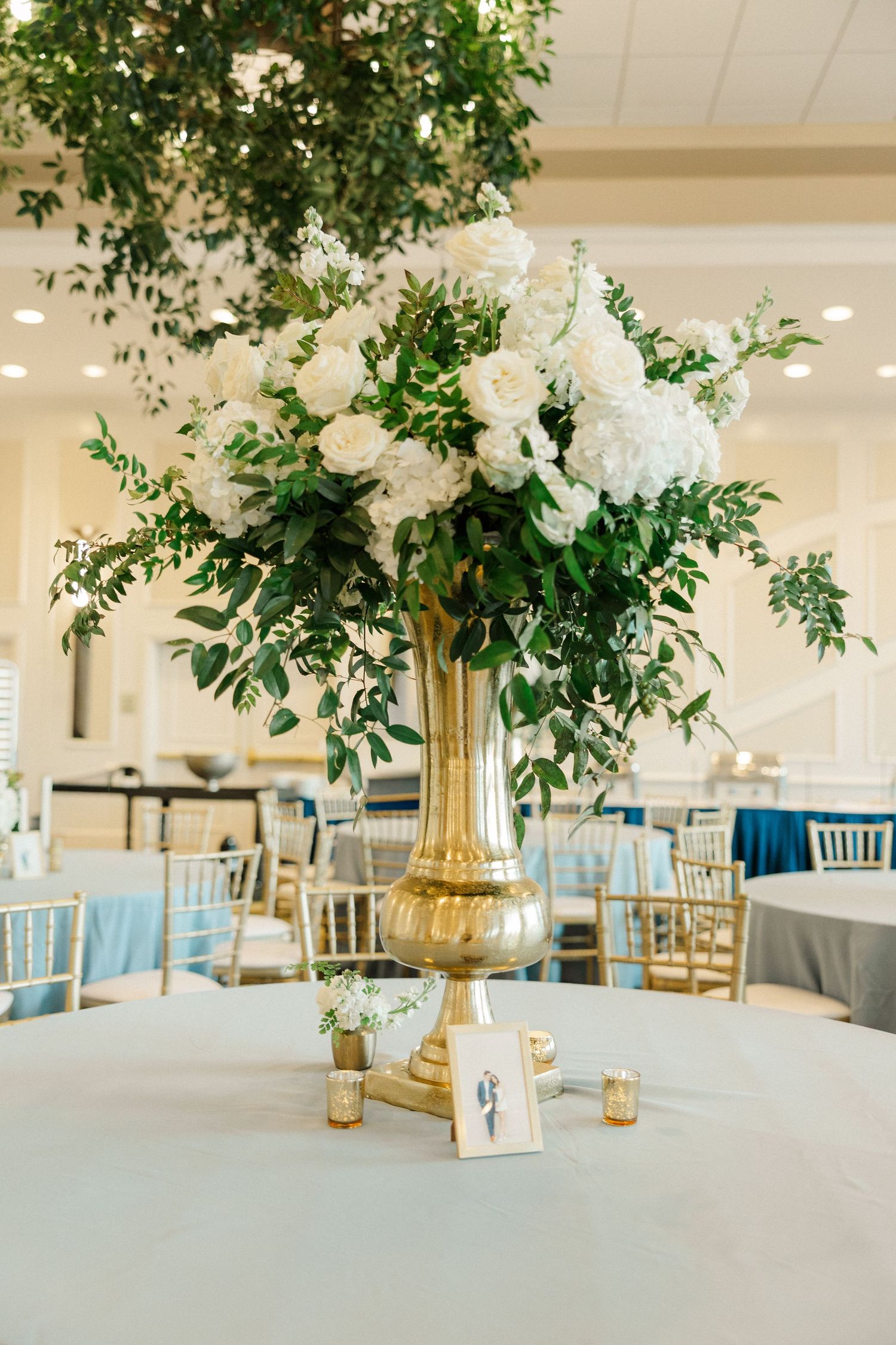 Tall gold vase centerpiece with white roses and lush greenery on a white tablecloth at an elegant wedding reception.