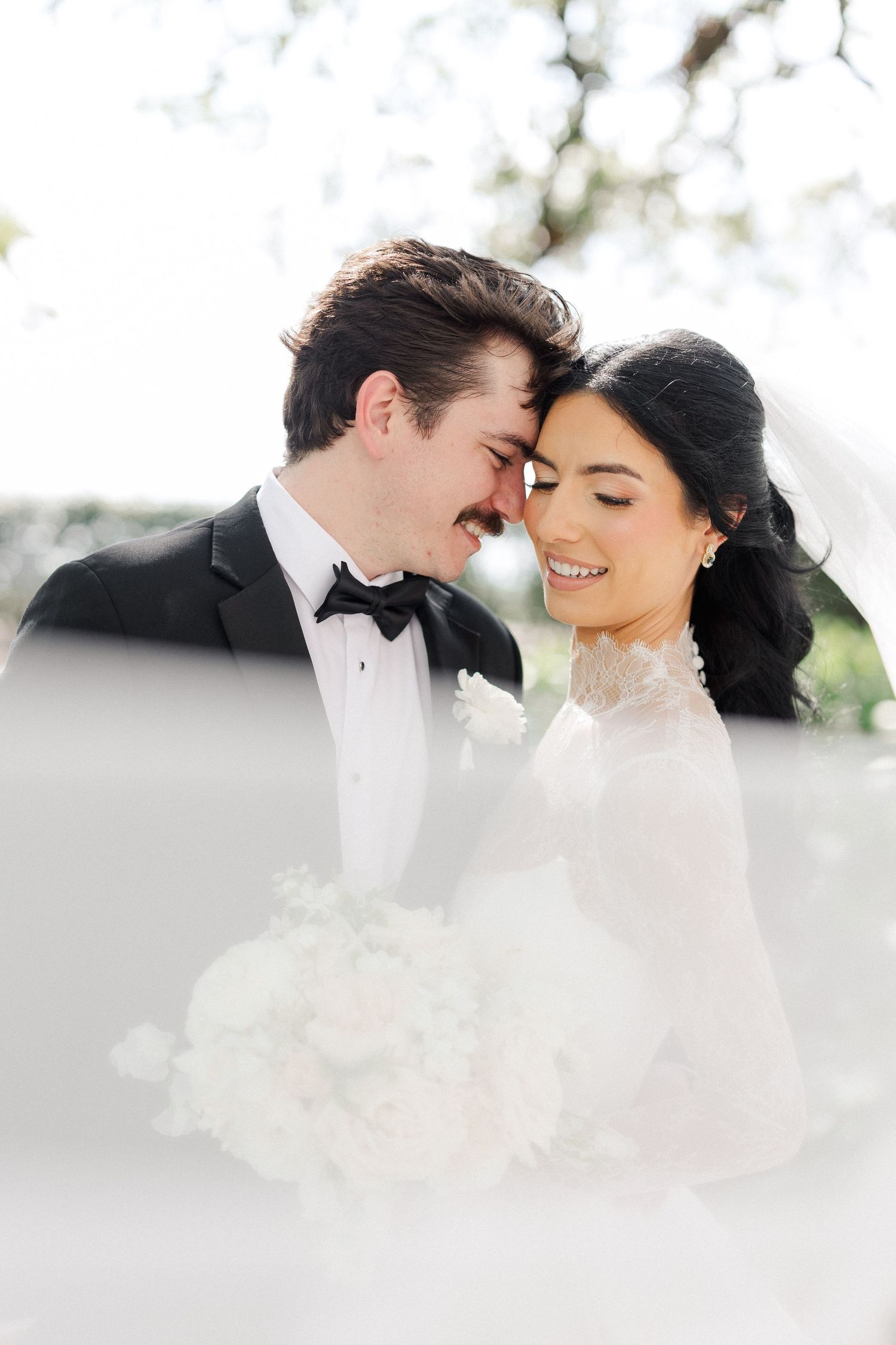 Bride and groom touch foreheads romantically outdoors, bride holding white bouquet in lace wedding dress.