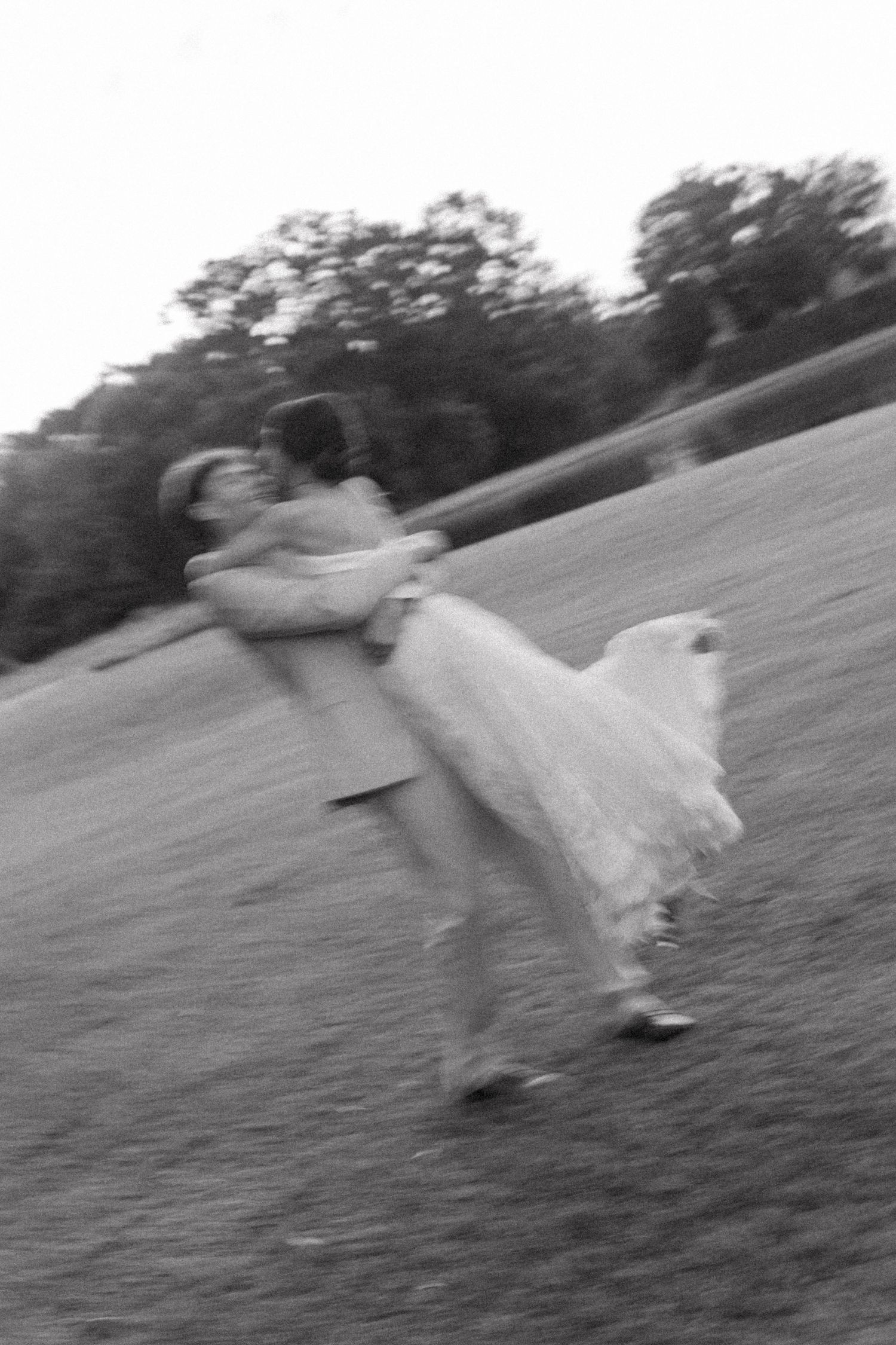 Black and white motion blur photo of groom lifting bride in wedding attire on an open grassy field.