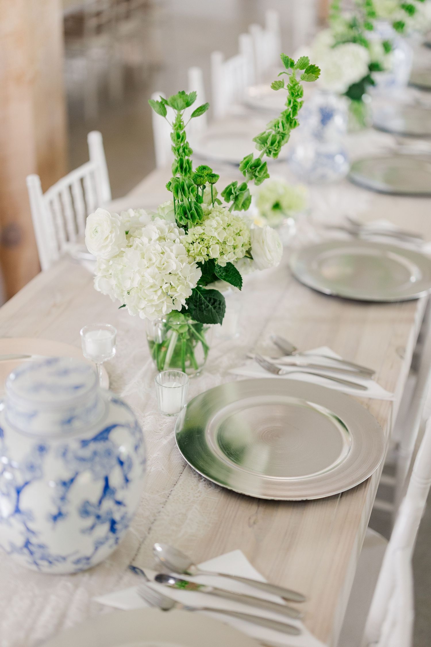 Elegant wedding reception table with silver charger plates, white hydrangeas, green florals, and blue and white ceramic vases.