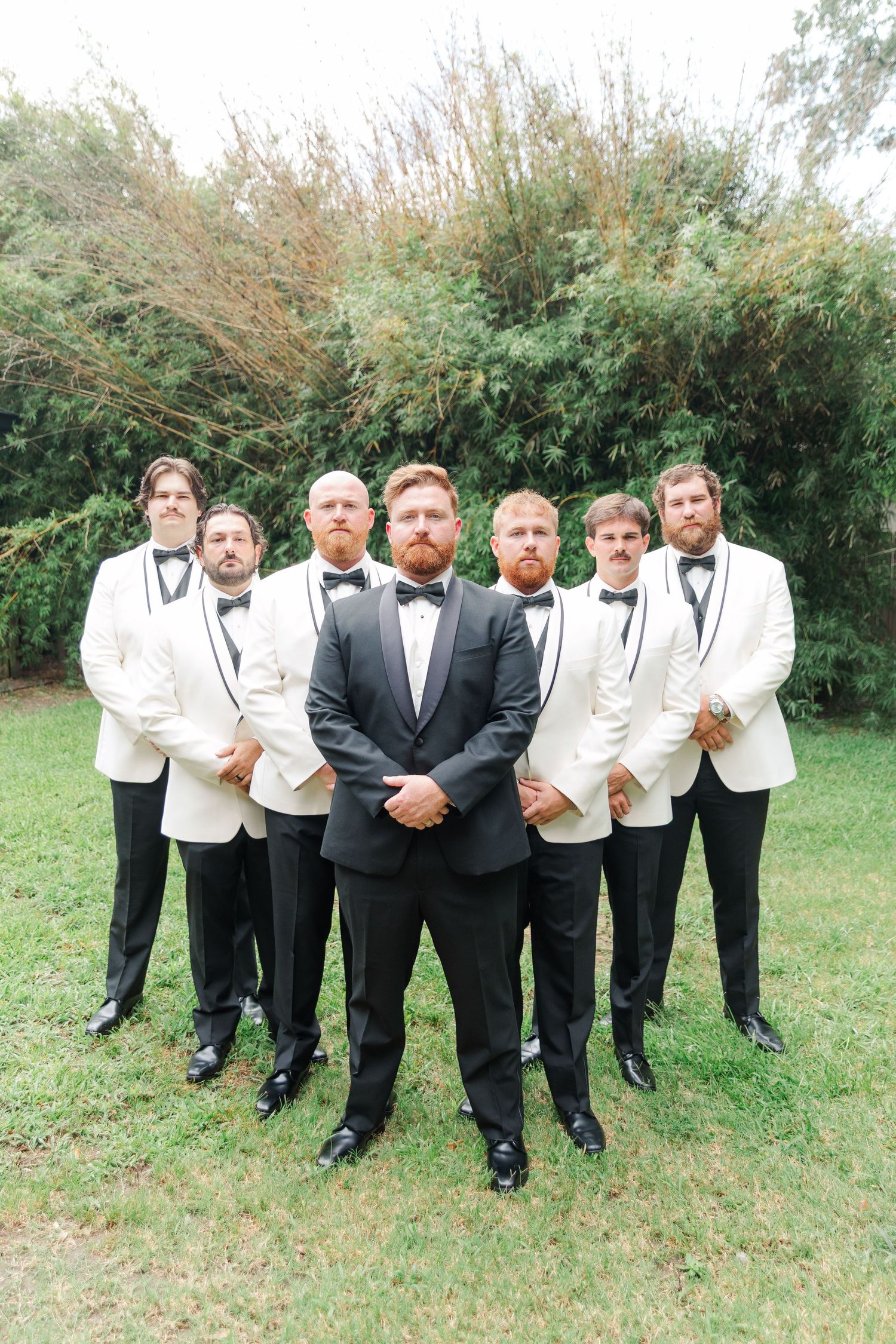 Groomsmen in formal attire striking fun poses for photos on a grassy lawn.
