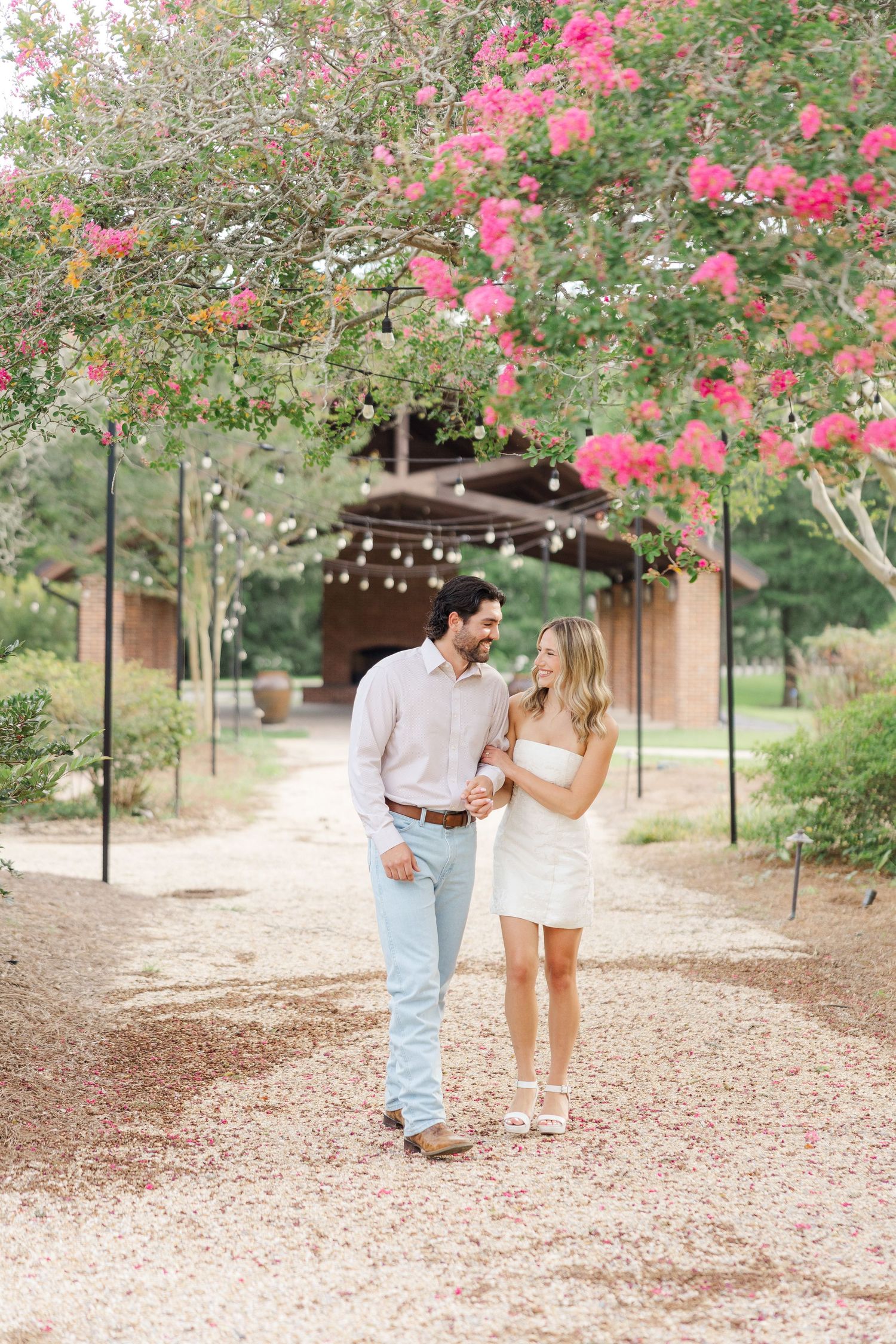 A couple walks along a path under blooming pink crepe myrtle trees on a summer day.