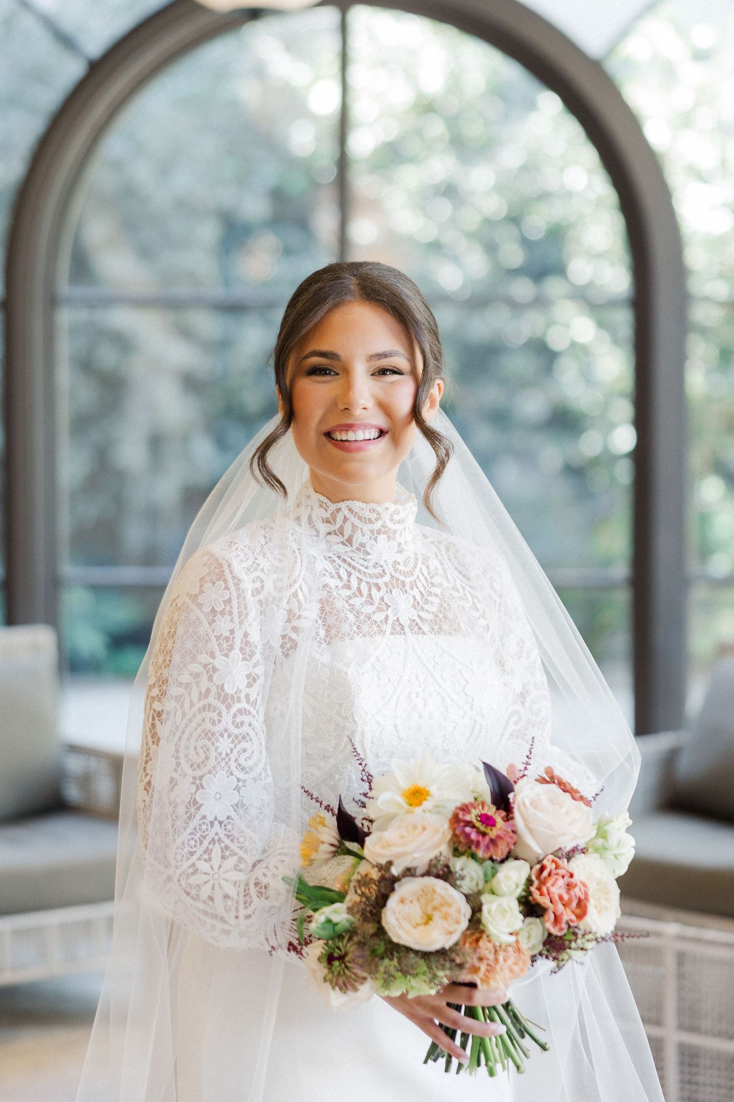 A bridal portrait showing a delicate lace gown and colorful spring bouquet in an arched window setting.