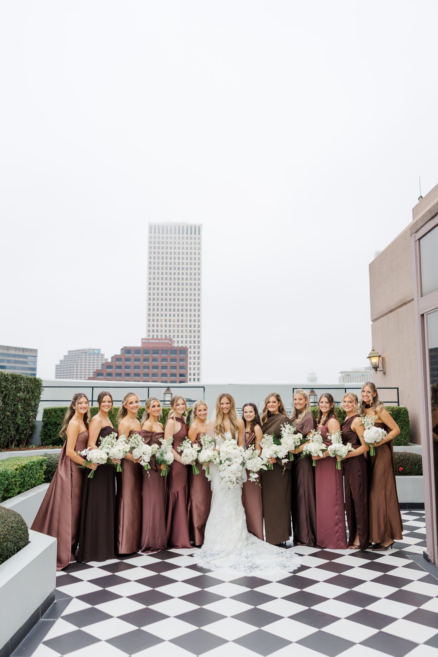 Wedding party in shades of brown dresses poses on checkered rooftop with city views.