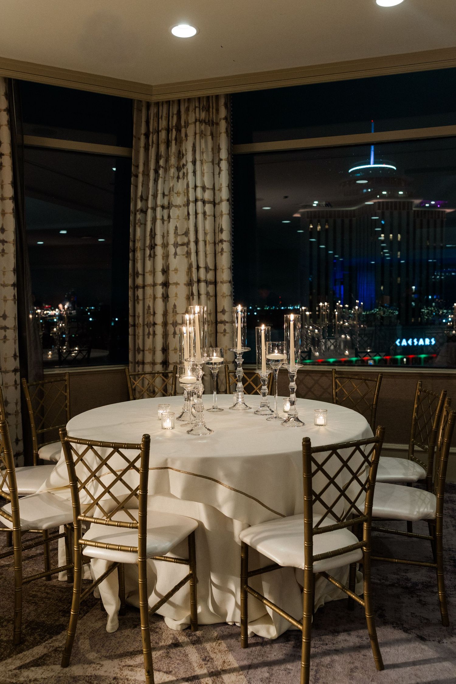An elegant reception venue with white tablecloths and gold chairs overlooking city lights at night.