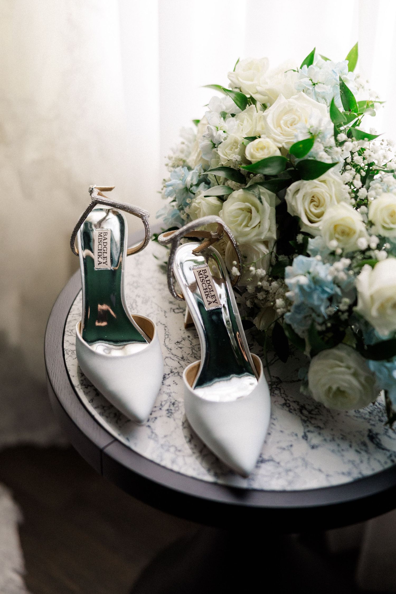 White bridal shoes displayed with elegant white roses and floral arrangements on a dark surface.