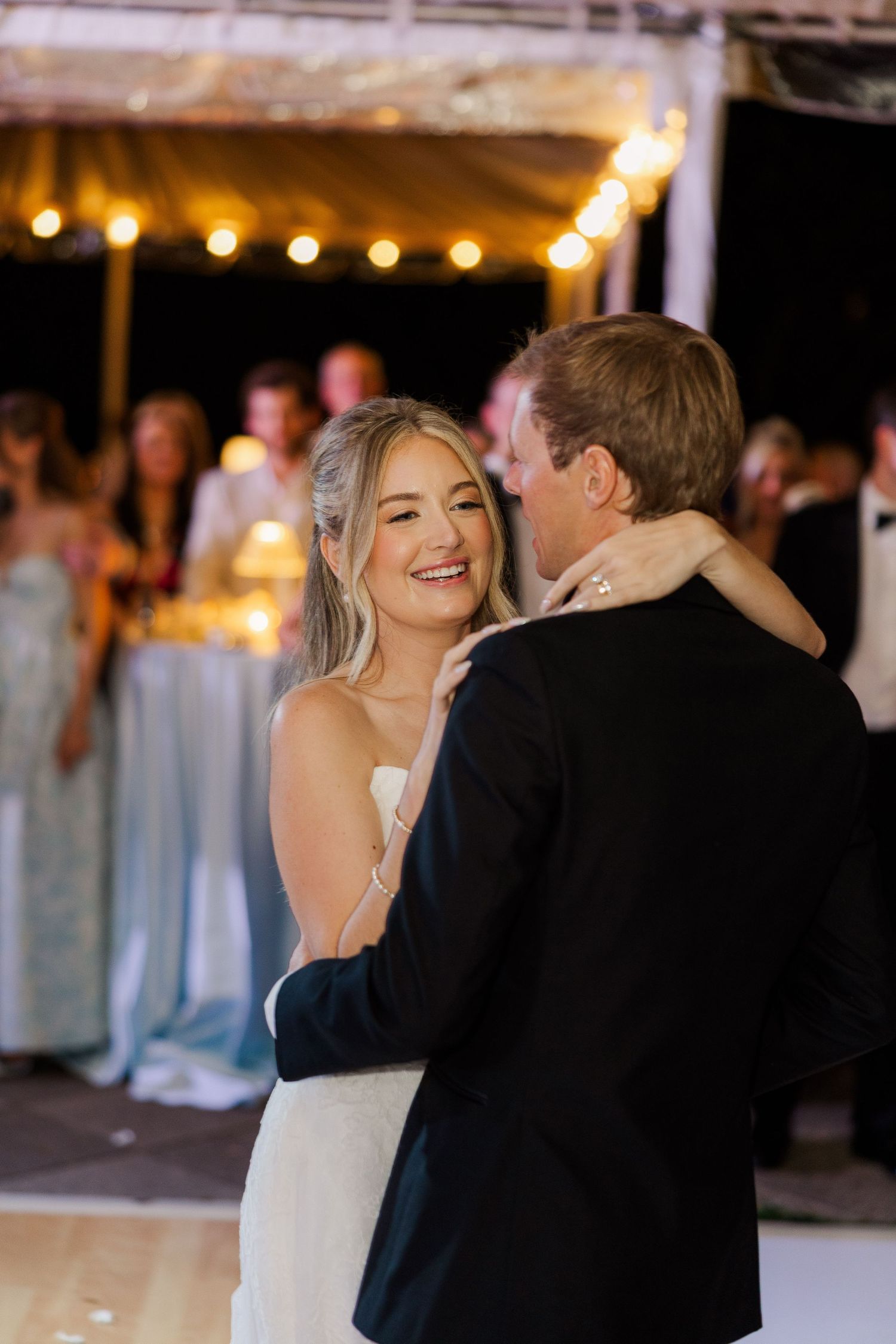 A couple shares a romantic dance moment under warm string lights at an outdoor evening wedding reception.