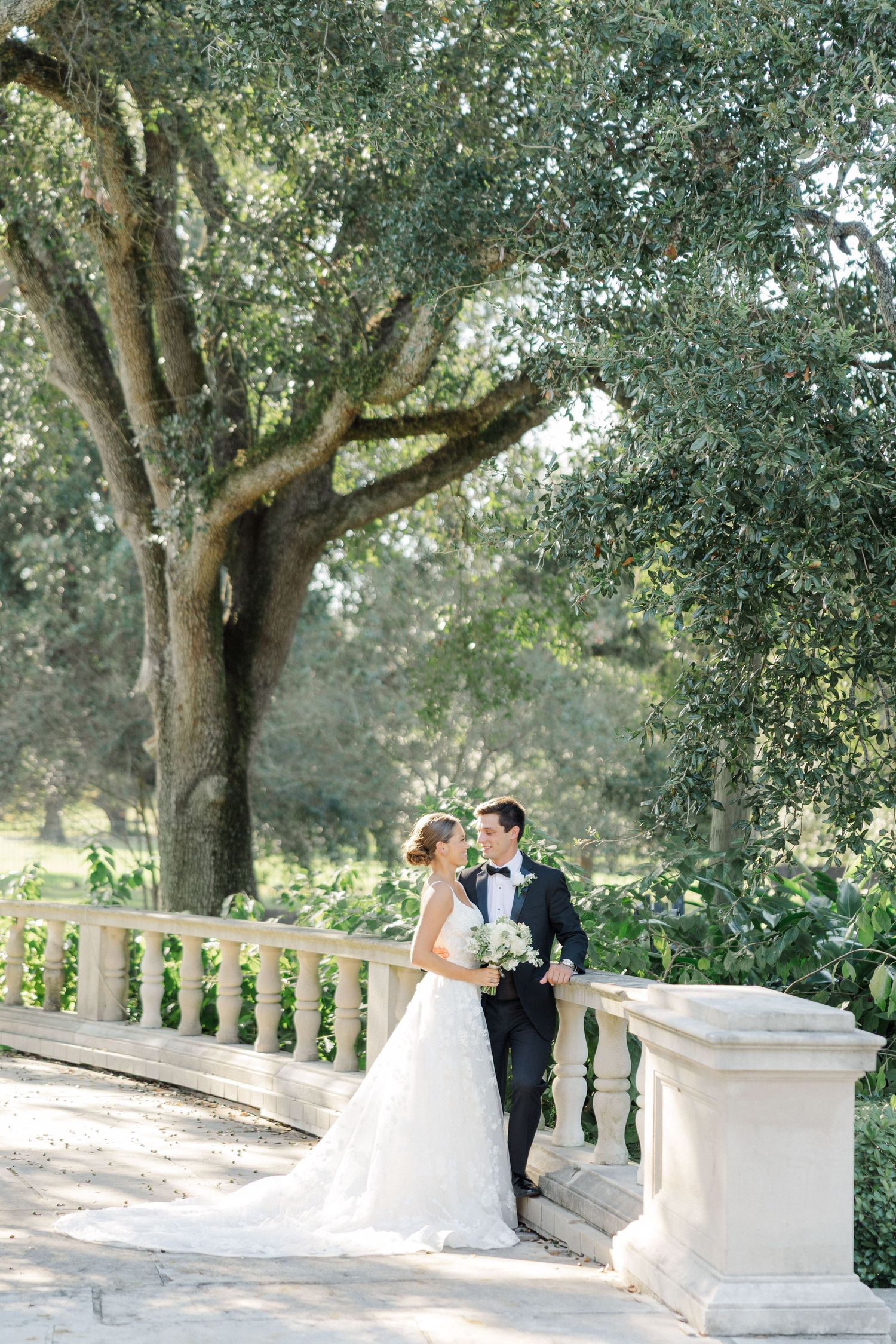 Wedding couple sharing an intimate moment under oak trees near a stone balustrade.