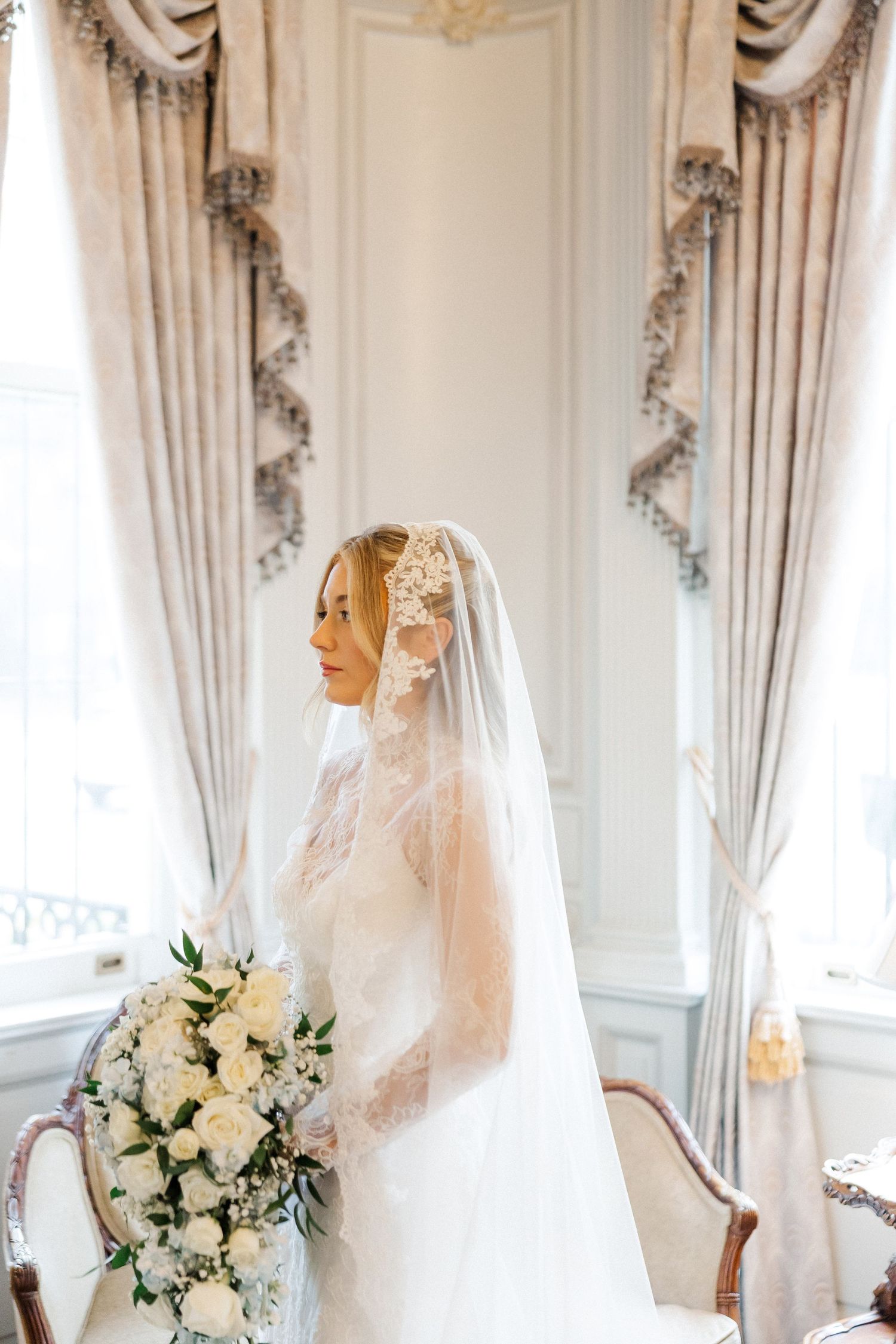 Elegant bridal portrait with white bouquet against ornate curtained window.