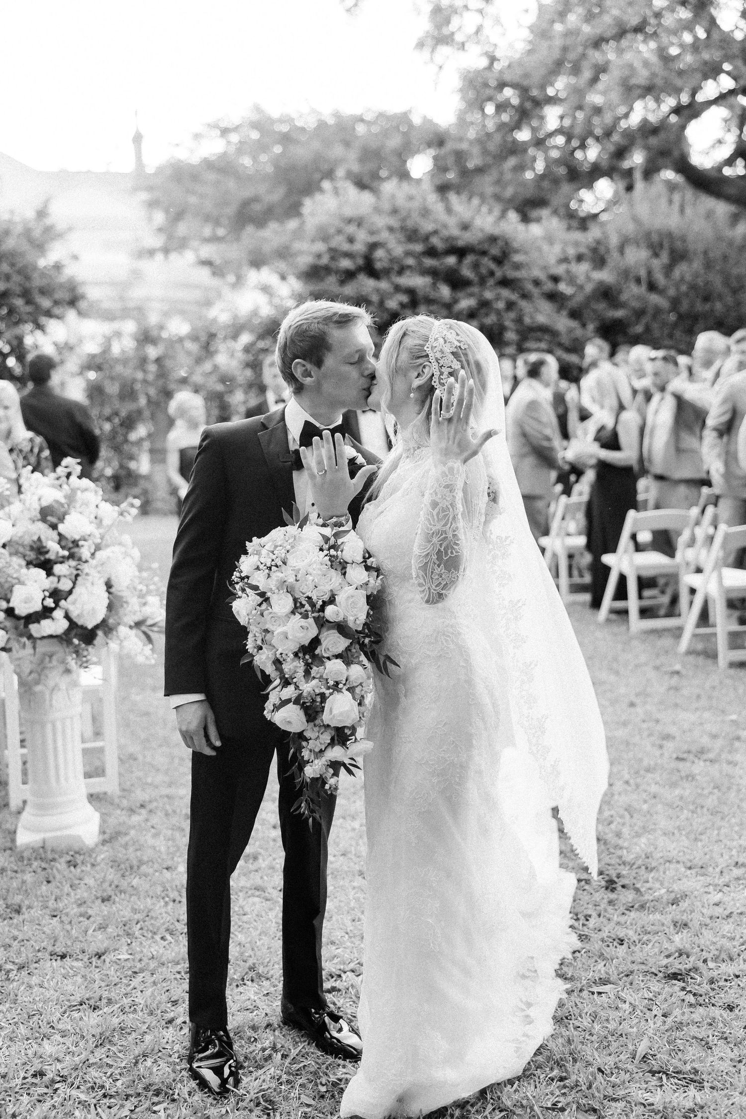 Outdoor wedding ceremony setup with white chairs and floral arrangements under oak trees.