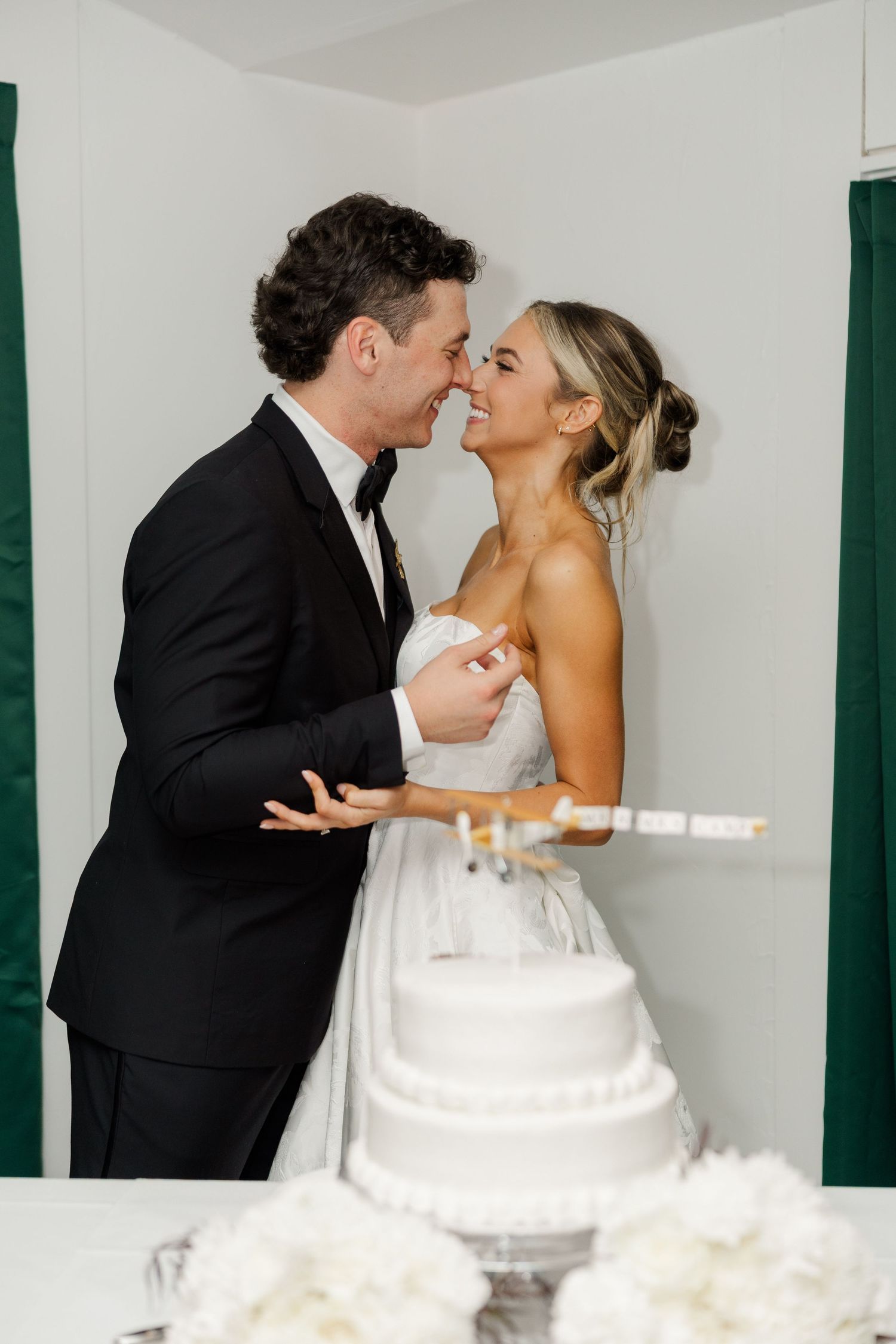 A wedding couple shares intimate moments beside their white tiered wedding cake at the reception.